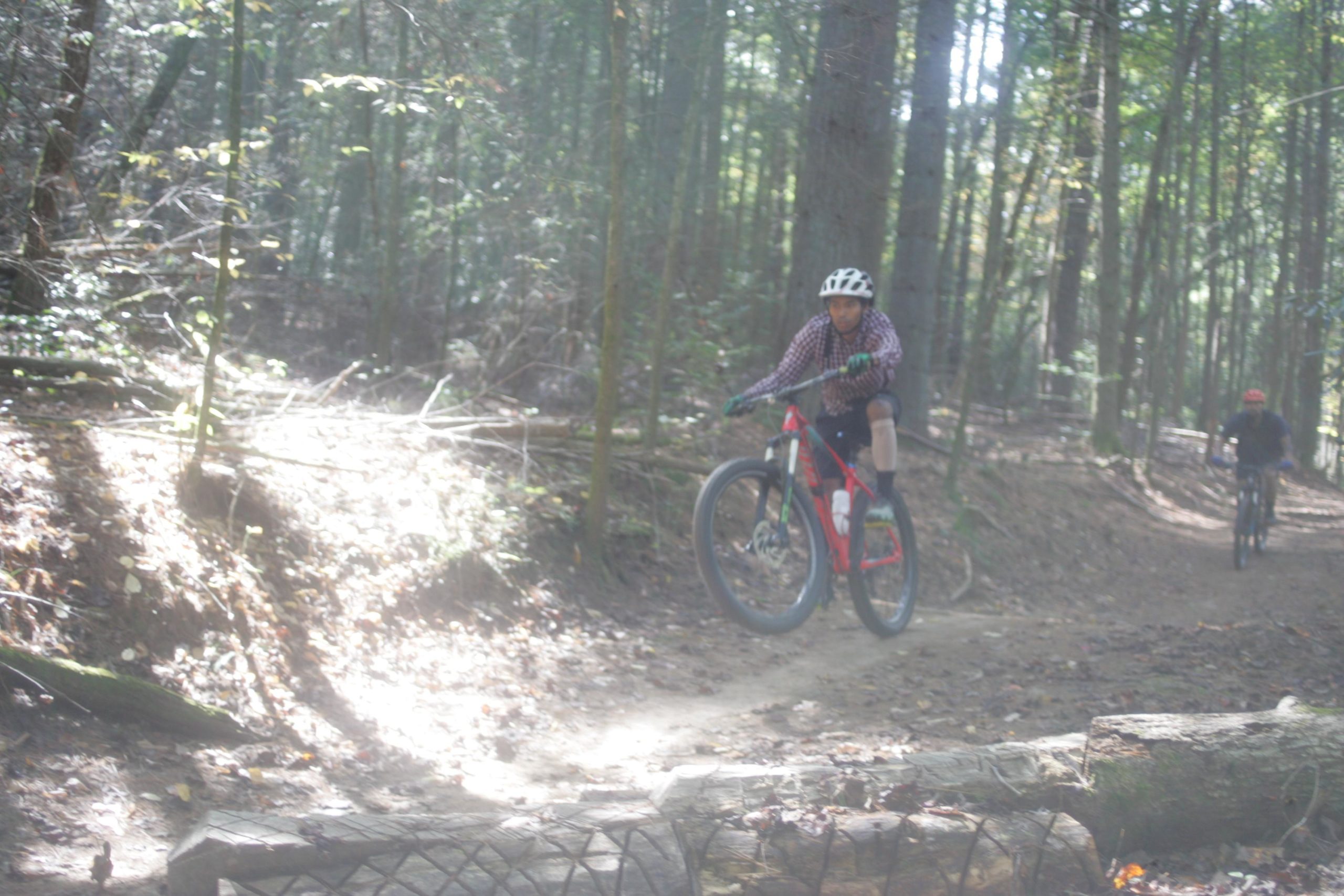 A mountain biker in a checkered shirt and helmet is performing a jump over a fallen log while riding on a dirt trail in a forest. Another biker is visible in the background, following the same trail. The scene captures the excitement of mountain biking in a natural setting with dappled sunlight filtering through the trees. Bent Creek mountain bike trail.