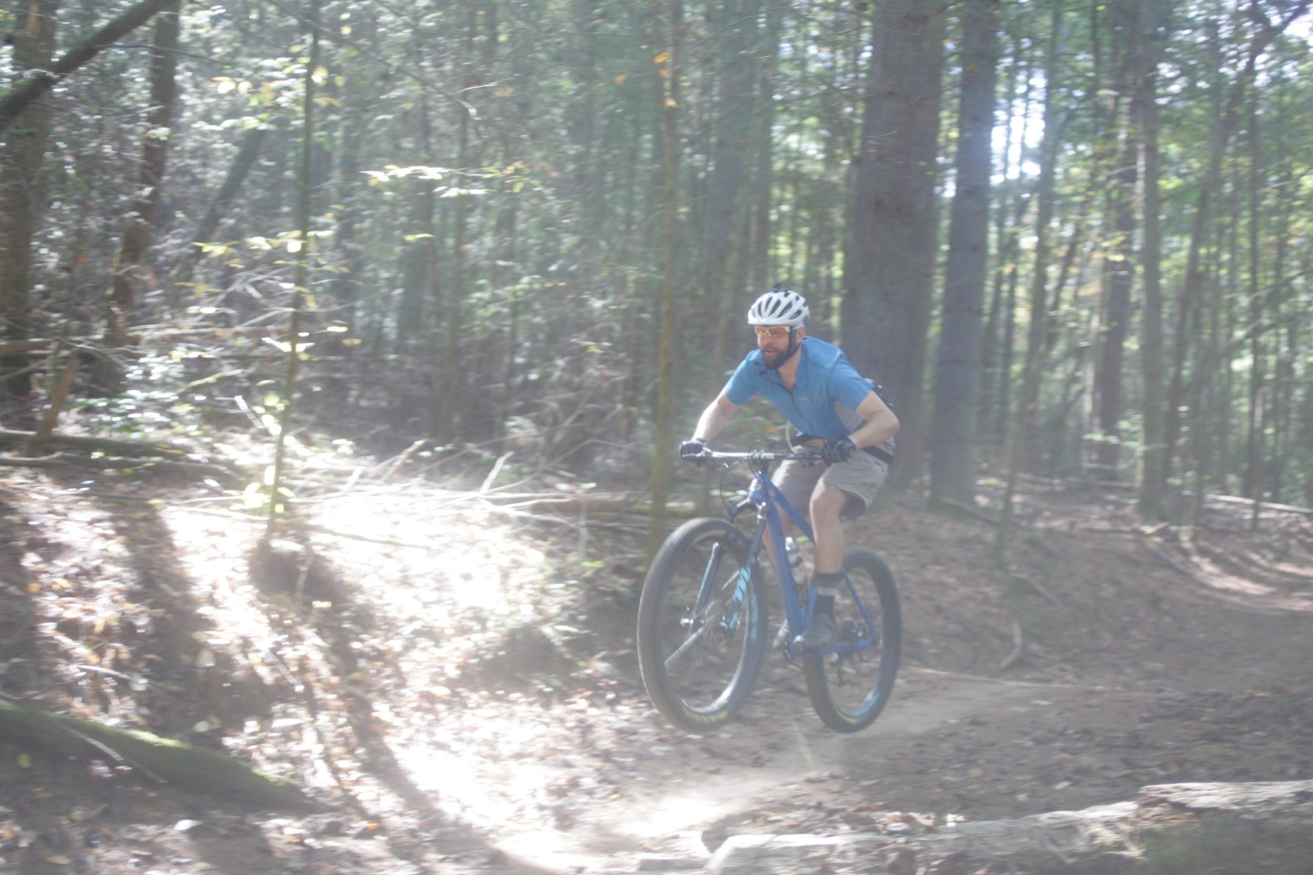 A cyclist in a blue shirt and helmet rides a mountain bike along a wooded trail, jumping over a small rise. Sunlight filters through the trees, casting dappled shadows on the ground while fallen leaves and branches dot the path. Bent Creek mountain bike trail.