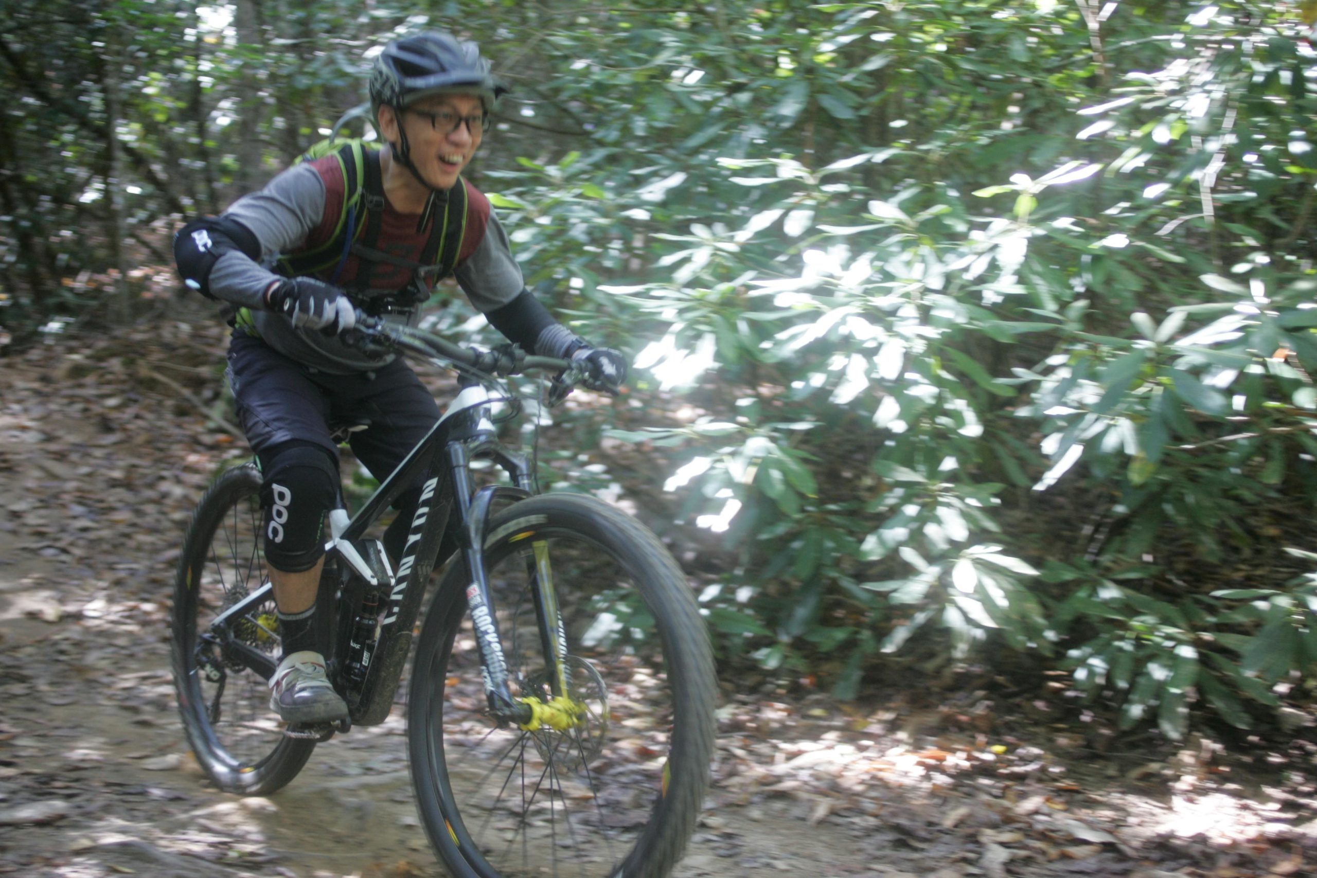 A mountain biker riding along a dirt trail surrounded by lush green foliage, wearing a helmet, protective gear, and a joyful expression. The bike features a black frame with yellow accents. Sunlight filters through the trees, creating a vibrant outdoor atmosphere. Bent Creek mountain bike trail.