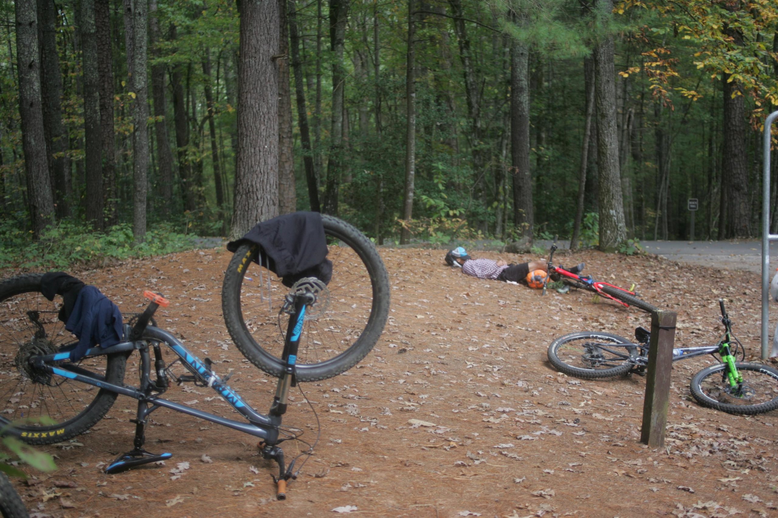 Two mountain bikes are placed on their sides on a dirt path surrounded by tall trees. One bike is leaning on its handlebars, while a second bike lies nearby. In the background, two individuals are lying on the ground; one is in a plaid shirt and the other is wearing a helmet and looking up. Fallen leaves cover the ground, indicating autumn. The scene conveys a relaxed outdoor atmosphere, possibly after a biking adventure. Bent Creek mountain bike trail.