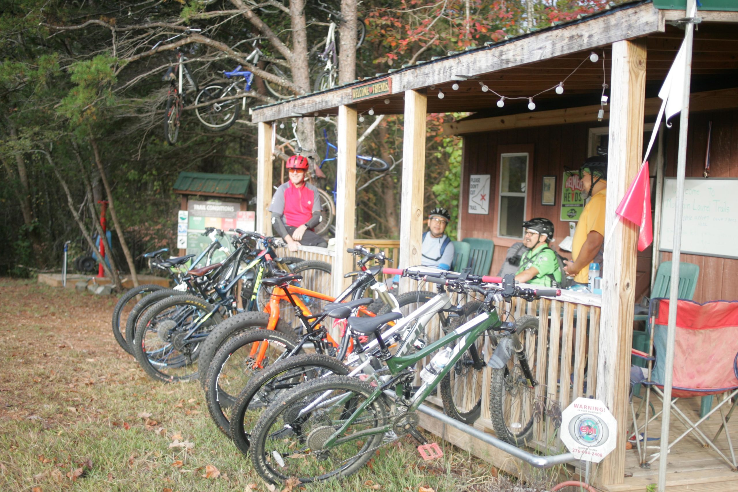 A group of mountain bikers is gathered at a wooden trailhead shelter. Several bicycles are parked in front of the shelter, with some bicycles hanging from a nearby tree. The riders are wearing helmets and cycling gear, and they are seated on the porch or standing near the shelter, which displays a sign welcoming visitors. The surrounding area is wooded, with some autumn foliage visible. Mountain Laurel Trails mountain bike trail.