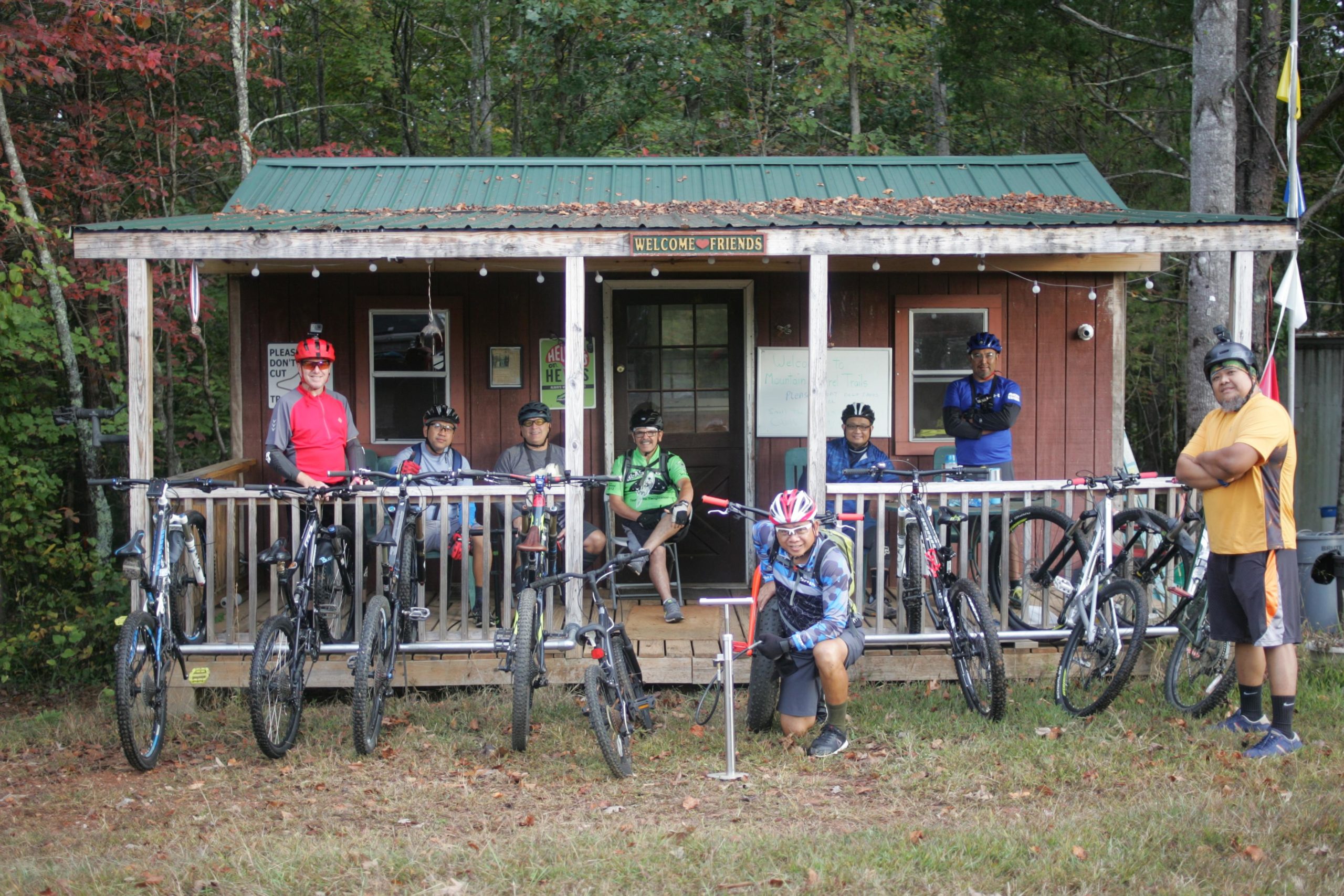 A group of seven mountain bikers posing in front of a cozy wooden cabin with a green metal roof. The cabin has a welcoming sign and is surrounded by lush trees with hints of autumn foliage. Six bikers are seated on the porch, while one is kneeling next to a bike pump. The cyclists are dressed in colorful biking gear and helmets, with bicycles lined up on either side. Mountain Laurel Trails mountain bike trail.