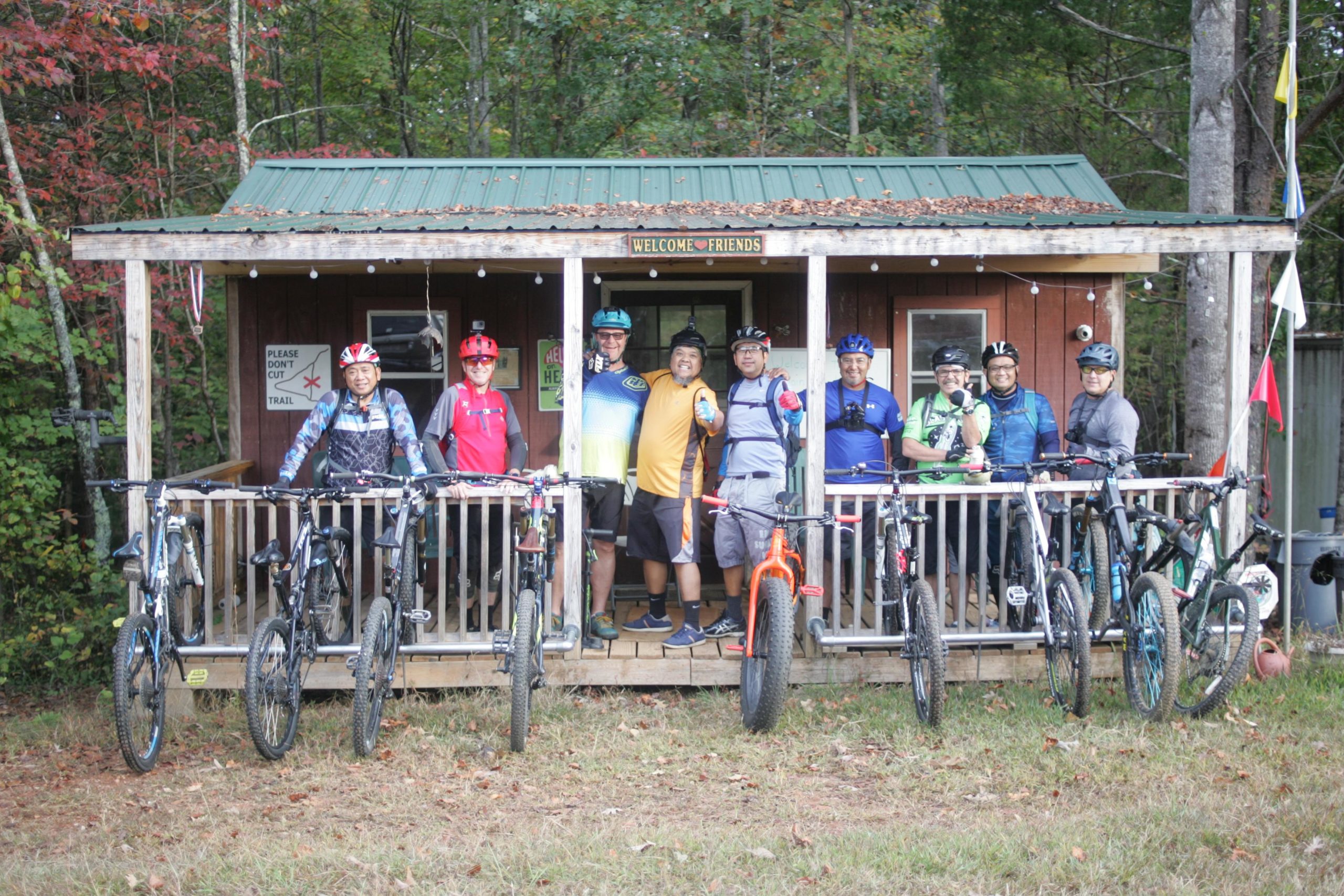 A group of eight mountain bikers pose together on the porch of a rustic wooden cabin, surrounded by trees with colorful autumn leaves. They are smiling and wearing cycling gear, with their bikes lined up beside them. The cabin has a green metal roof and signs that read "WELCOME FRIENDS" and "PLEASE DON'T CUT TRIAL." Mountain Laurel Trails mountain bike trail.