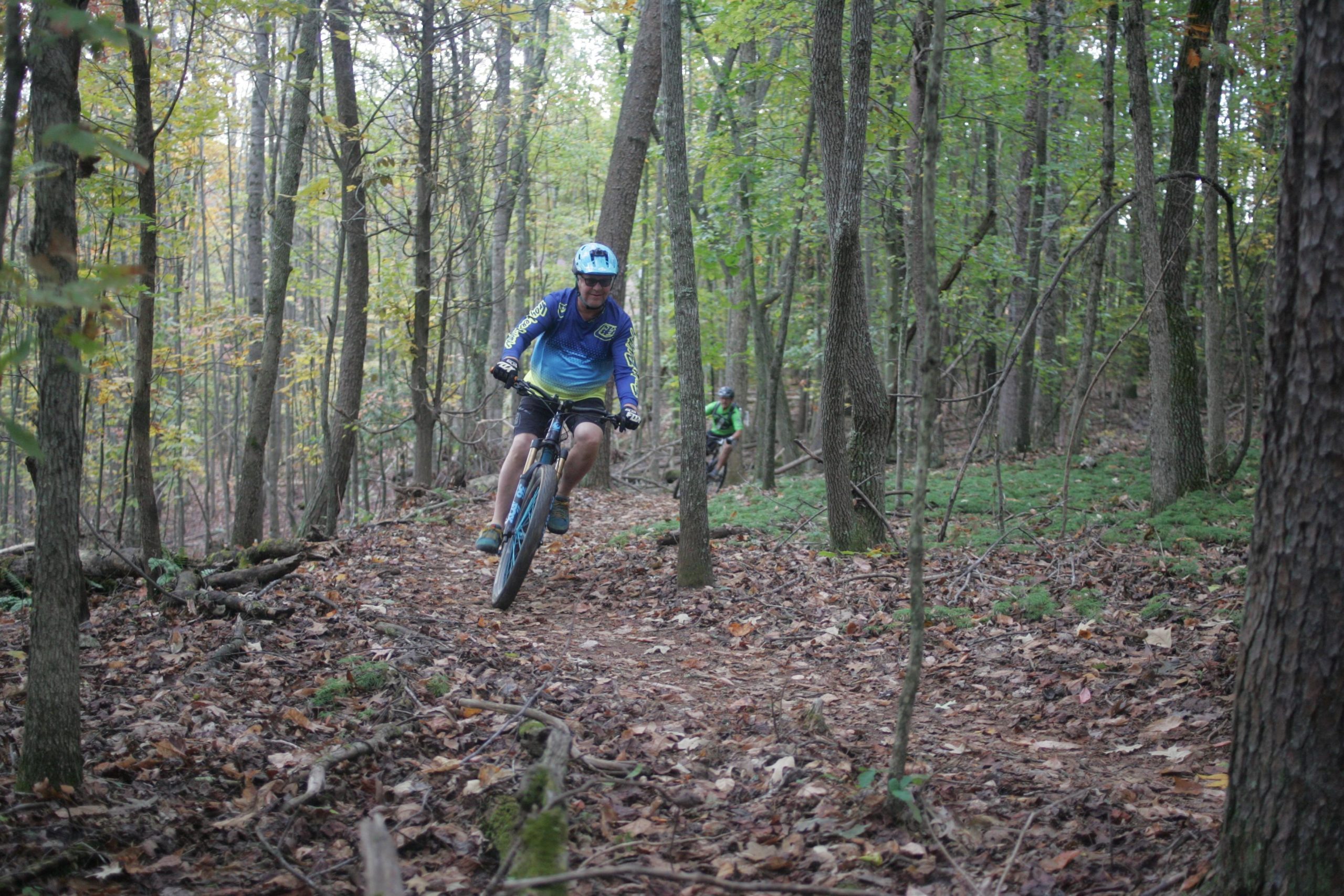 A person riding a mountain bike down a dirt trail surrounded by trees in a forested area. Another cyclist is visible in the background, also navigating the path. The scene captures an autumn atmosphere with leaves scattered on the ground. Mountain Laurel Trails mountain bike trail.