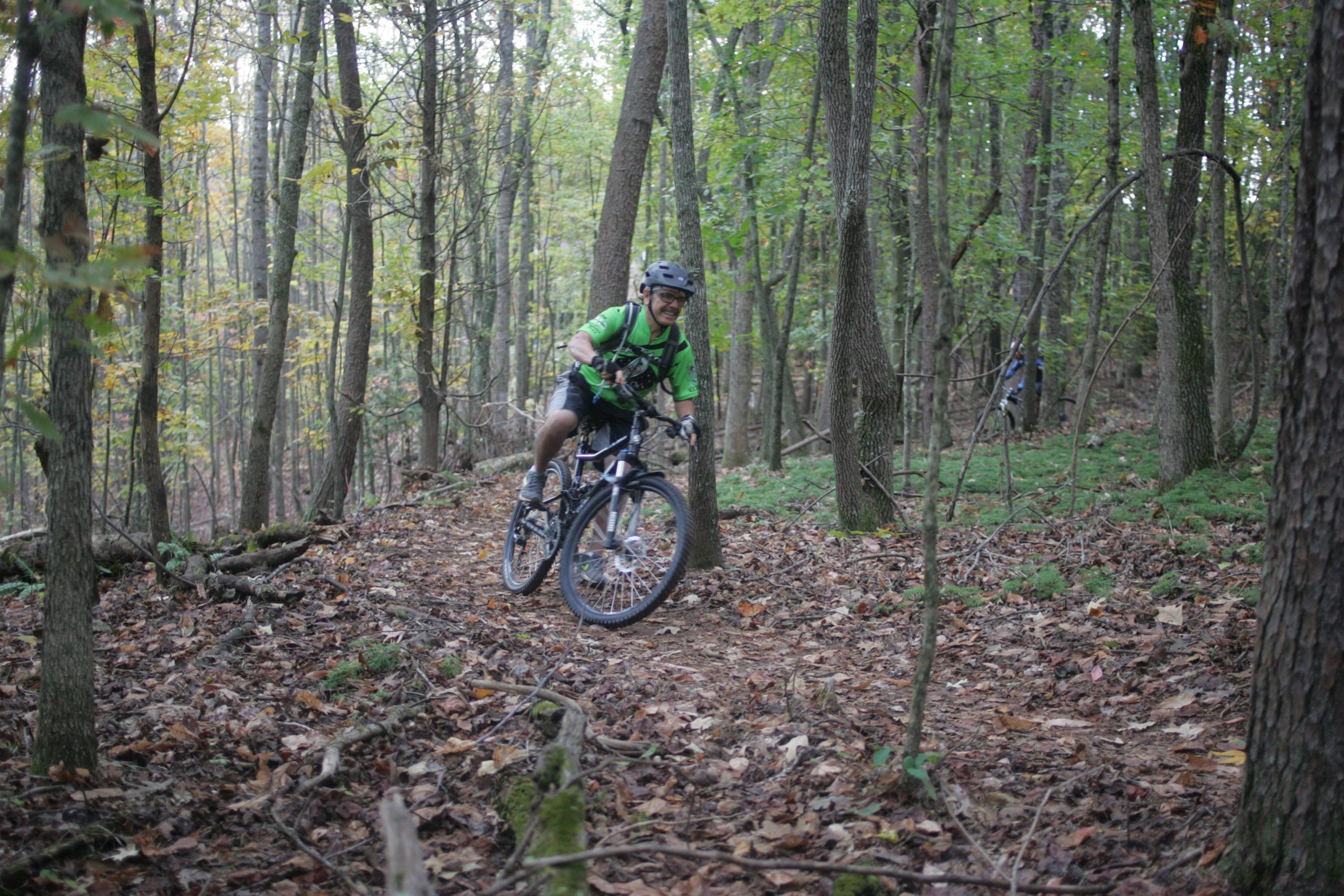 A mountain biker riding along a narrow, leaf-covered trail in a forest. The cyclist is wearing a green shirt and a helmet, and is leaning into the turn with a smile. Surrounding him are tall trees with green leaves and scattered autumn foliage on the ground. Mountain Laurel Trails mountain bike trail.