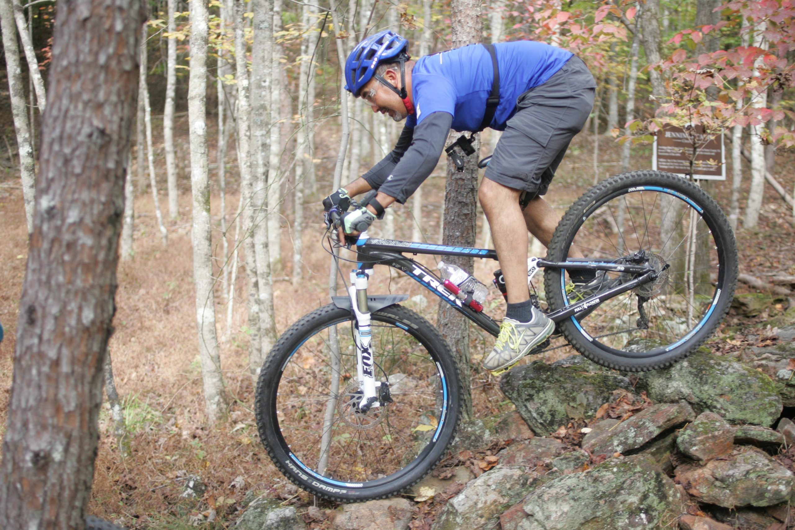 A mountain biker navigates rocky terrain in a forested area, wearing a blue helmet and shirt, with a focused expression as he balances on his bicycle. The scene features trees with autumn foliage in the background, showcasing a vibrant natural setting. Mountain Laurel Trails mountain bike trail.