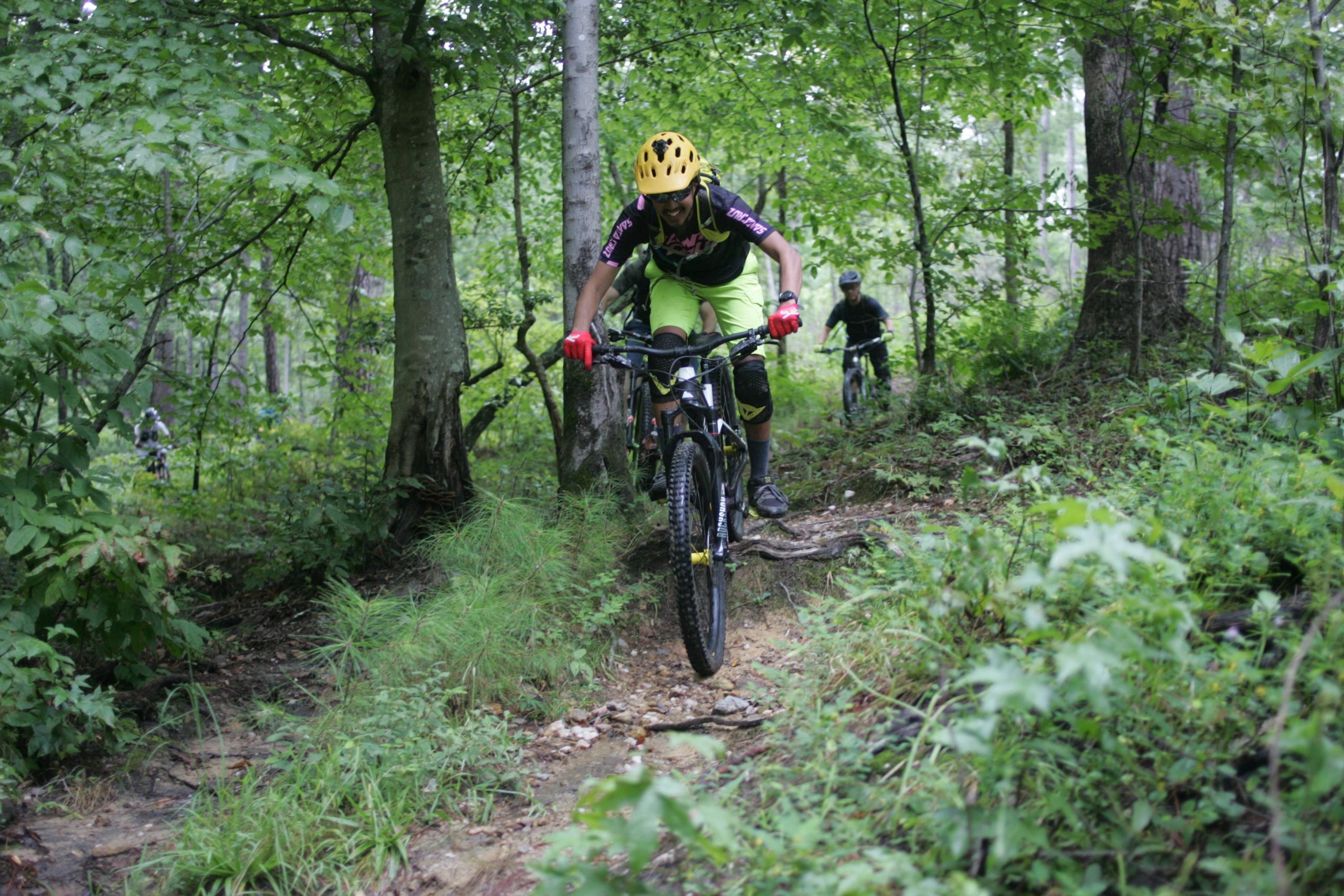 A mountain biker navigating a rocky trail surrounded by lush green trees and foliage, wearing a helmet and protective gear. Another biker can be seen in the background, also riding along the path. New Light Trails mountain bike trail.