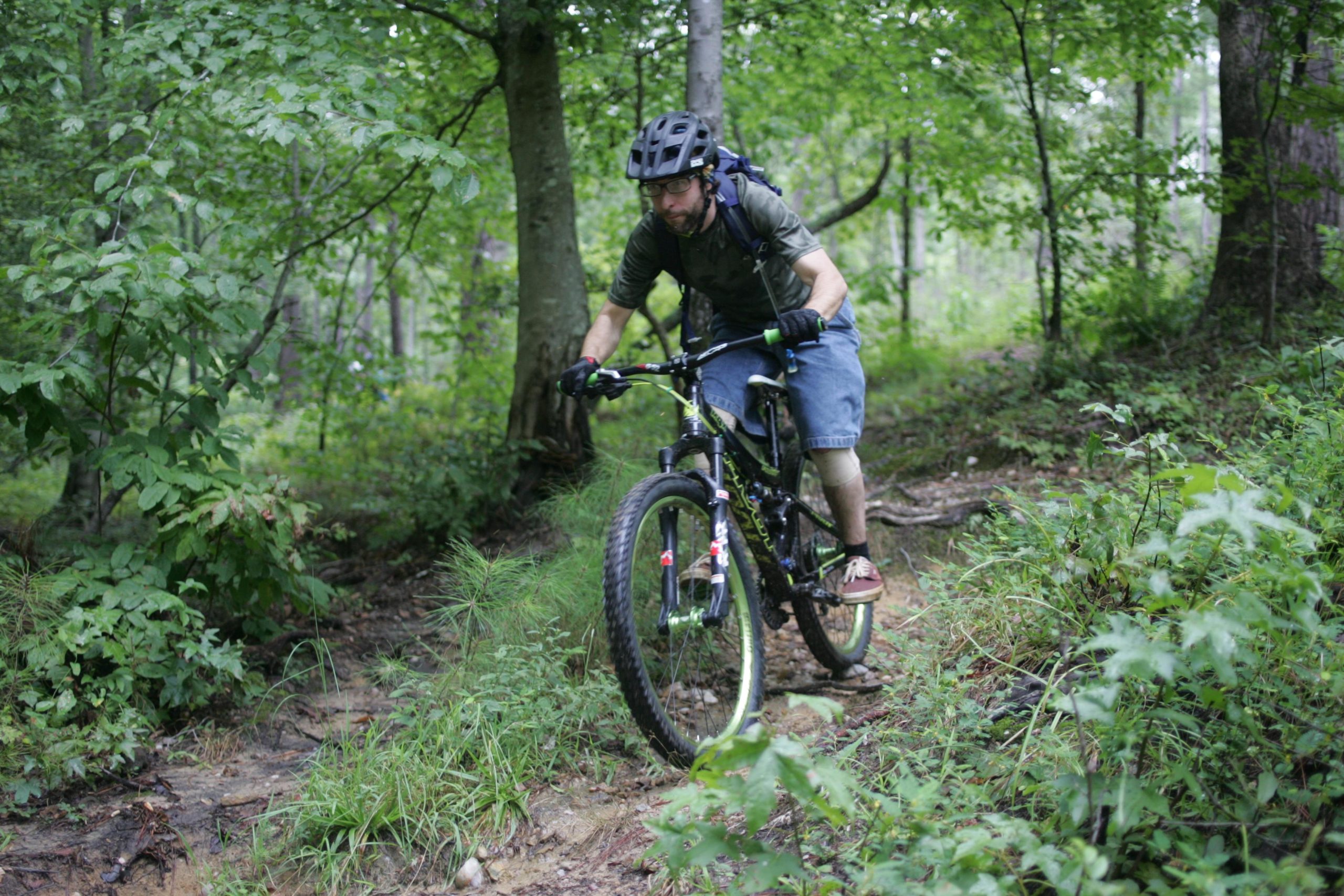 A person riding a mountain bike on a narrow, dirt trail surrounded by lush greenery in a forested area. The rider is wearing a black helmet, gloves, and a casual outfit consisting of a shirt and shorts, with a backpack visible on their back. New Light Trails mountain bike trail.