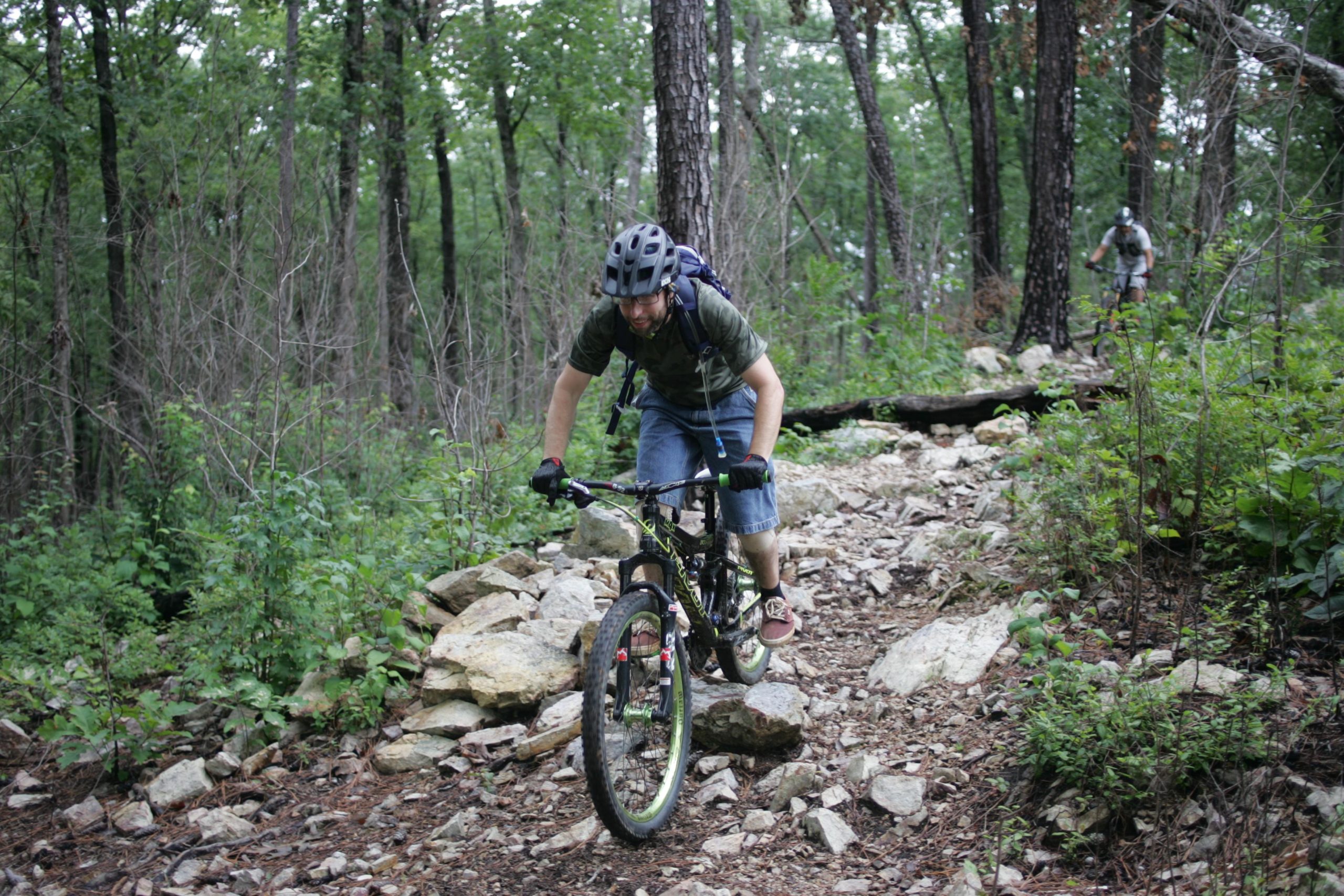A mountain biker navigates a rocky trail through a lush, green forest, focused on balancing and maneuvering their bike over the terrain. Another cyclist is visible in the background, also riding along the trail. New Light Trails mountain bike trail.
