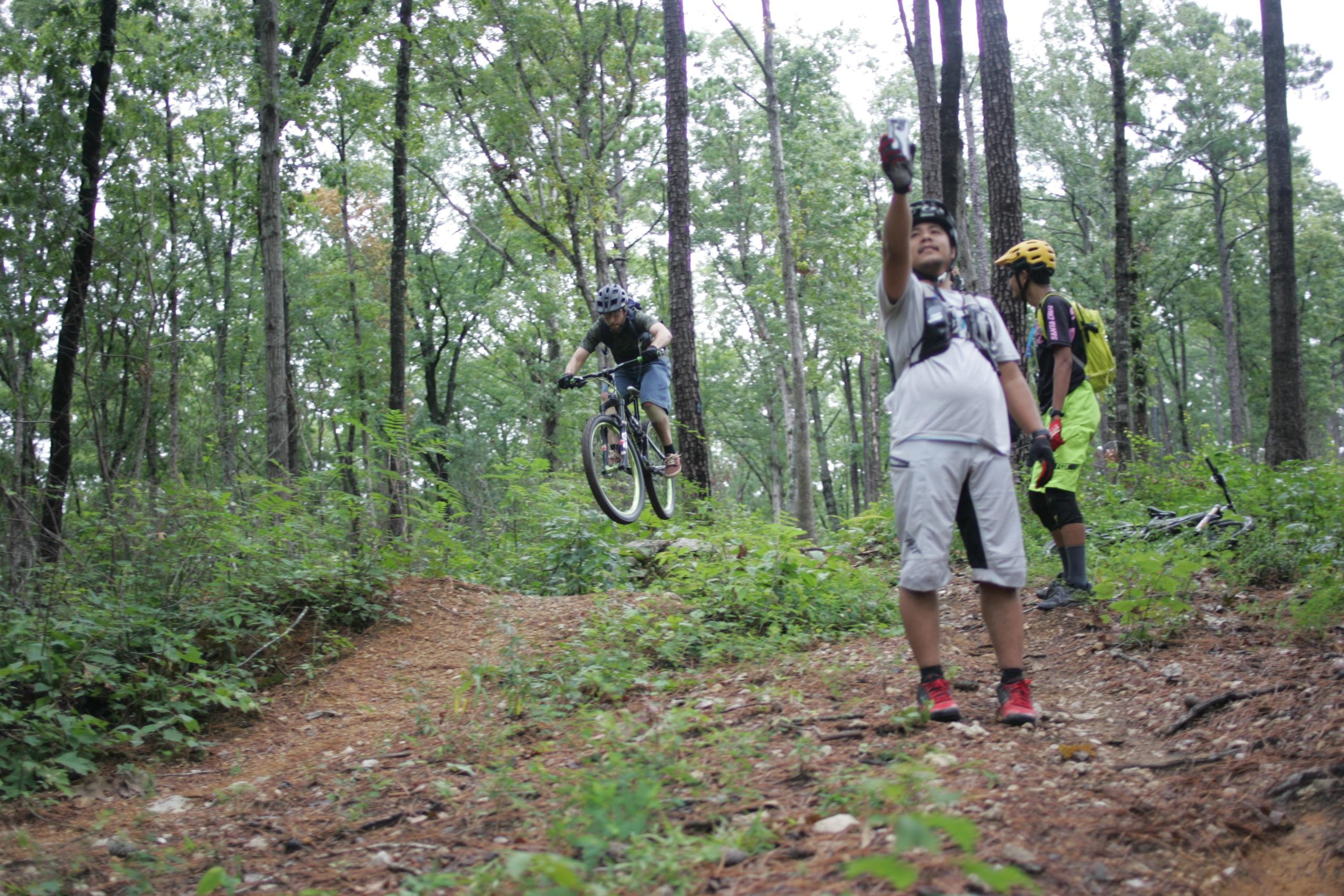 A mountain biker performing a jump on a trail in a wooded area, with two other bikers in the foreground; one person is pointing and observing while the other stands behind. The scene is surrounded by green foliage and tall trees, creating a lively outdoor atmosphere. New Light Trails mountain bike trail.