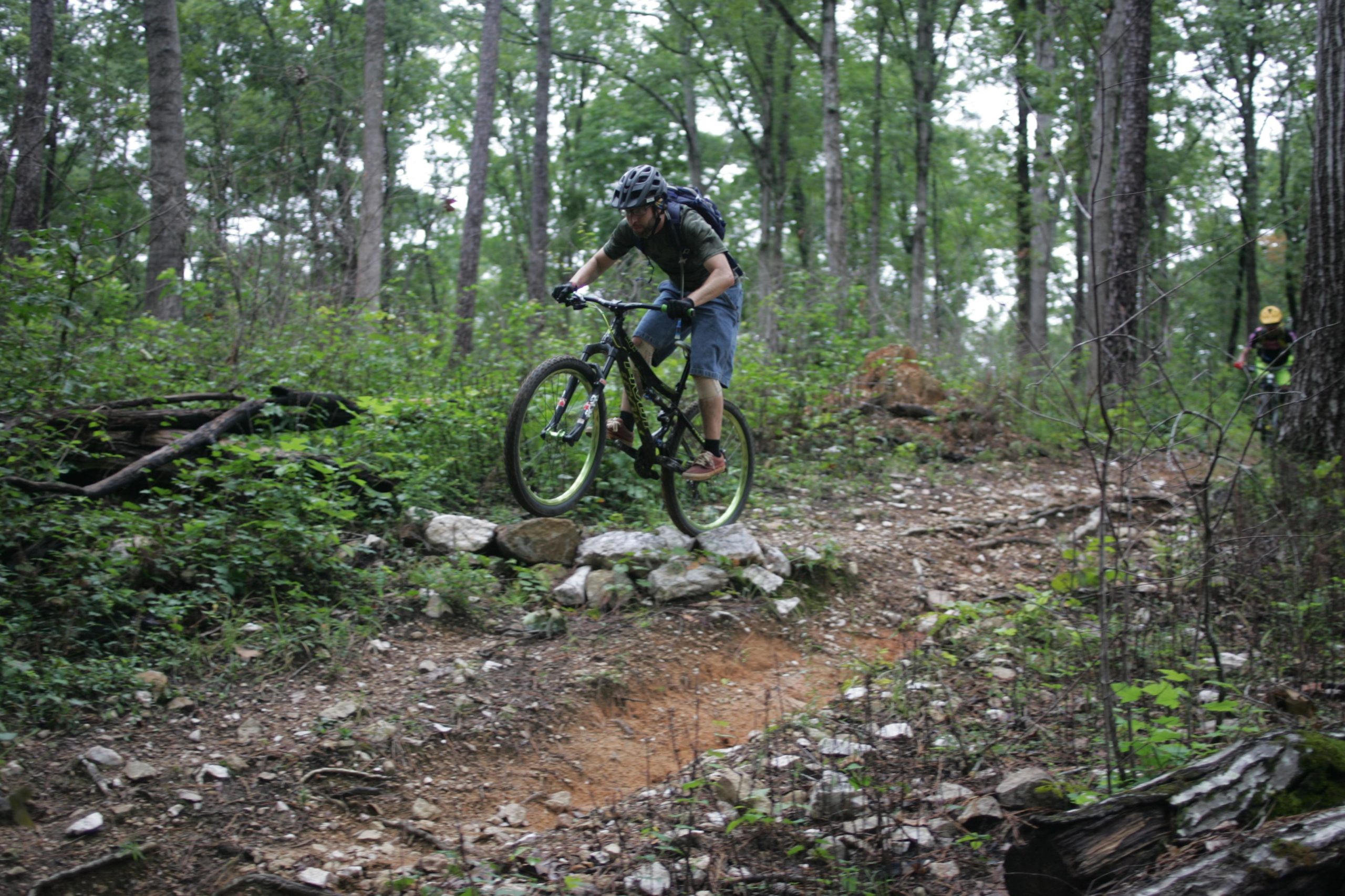 A mountain biker performing a jump over rocky terrain in a wooded area, surrounded by tall trees and lush greenery. New Light Trails mountain bike trail.