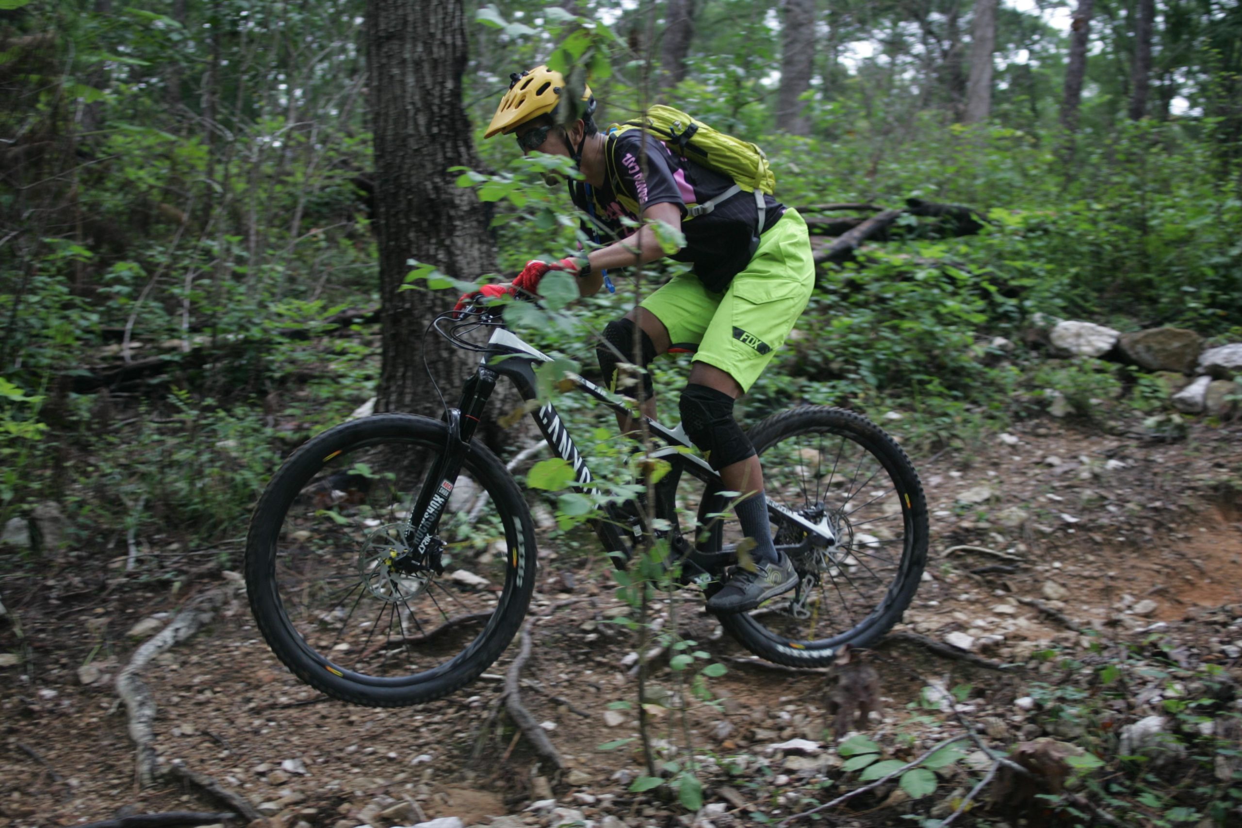 A mountain biker in bright yellow shorts and a helmet navigates a rugged, overgrown trail surrounded by trees and rocks, caught mid-jump as they ride over a rocky terrain. New Light Trails mountain bike trail.