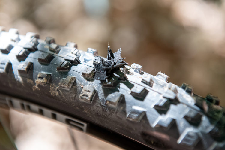 Close-up view of a mountain bike tire showing a section with worn-out rubber and a small piece of debris embedded in the tread. The tire has a rugged design suitable for off-road use, with dirt visible on its surface.