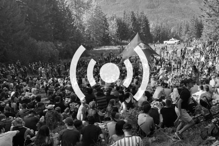 A large crowd of people seated on grass, gathered at an outdoor event in a forested area. A colorful tent is visible in the background, and a vehicle can be seen further back. The image has a graphic overlay representing sound waves, suggesting a musical or performance event.