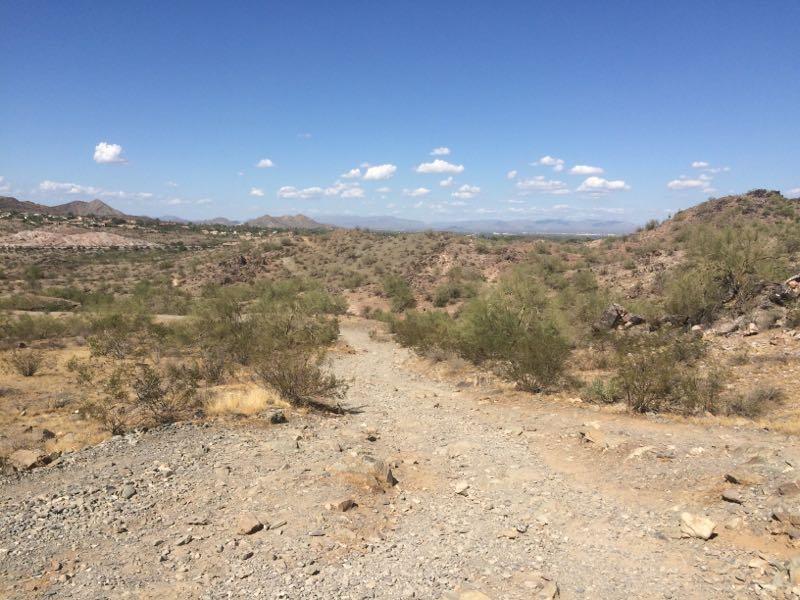 A dirt path winding through a dry, rugged landscape with sparse vegetation, surrounded by rocky hills under a clear blue sky with a few white clouds. Charles M. Christiansen (100) mountain bike trail.