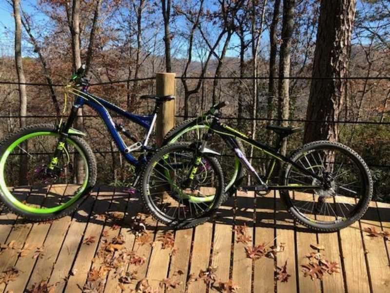 A blue and green mountain bike and a black and green mountain bike are parked on a wooden deck surrounded by trees in a forest setting. The background features autumn foliage and a clear blue sky. Fire Mountain Trail System mountain bike trail.