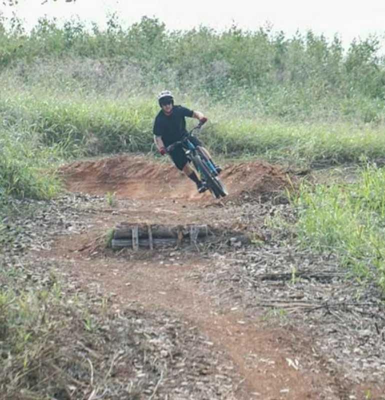 A mountain biker wearing a helmet and black clothing leans into a turn on a dirt trail, navigating a jump made of logs in a green, grassy outdoor setting. 5 Eighty Trails mountain bike trail.