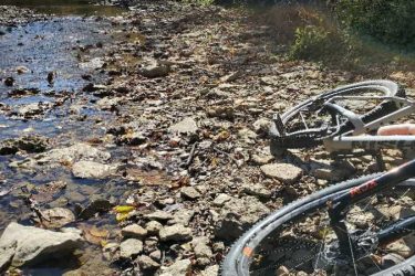 A close-up view of two mountain bikes resting on rocky terrain near a shallow stream, surrounded by fallen leaves and vegetation. The sunlight casts a warm glow over the scene, highlighting the natural beauty of the outdoor surroundings. Versailles State Park mountain bike trail.