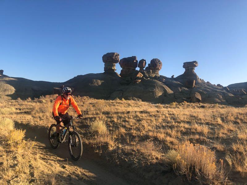 A mountain biker in an orange jacket rides along a dirt trail, with unique rock formations in the background under a clear blue sky. The terrain is dry, featuring golden grasses and rocky outcrops. Double Slot Fat Bike Trail mountain bike trail.