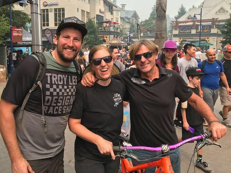 Commencal Meta AM V4: A group of three smiling individuals poses together outdoors in a busy setting. The background shows a crowd of people and buildings, with a slight haze in the air. One person on the left, wearing a black and gray checked shirt, stands next to a woman in sunglasses and a black shirt, who has her arms around him and smiles brightly. On the right, a man with sunglasses and longer hair holds a bicycle with a pink handlebar. They all appear happy and engaged in their surroundings.