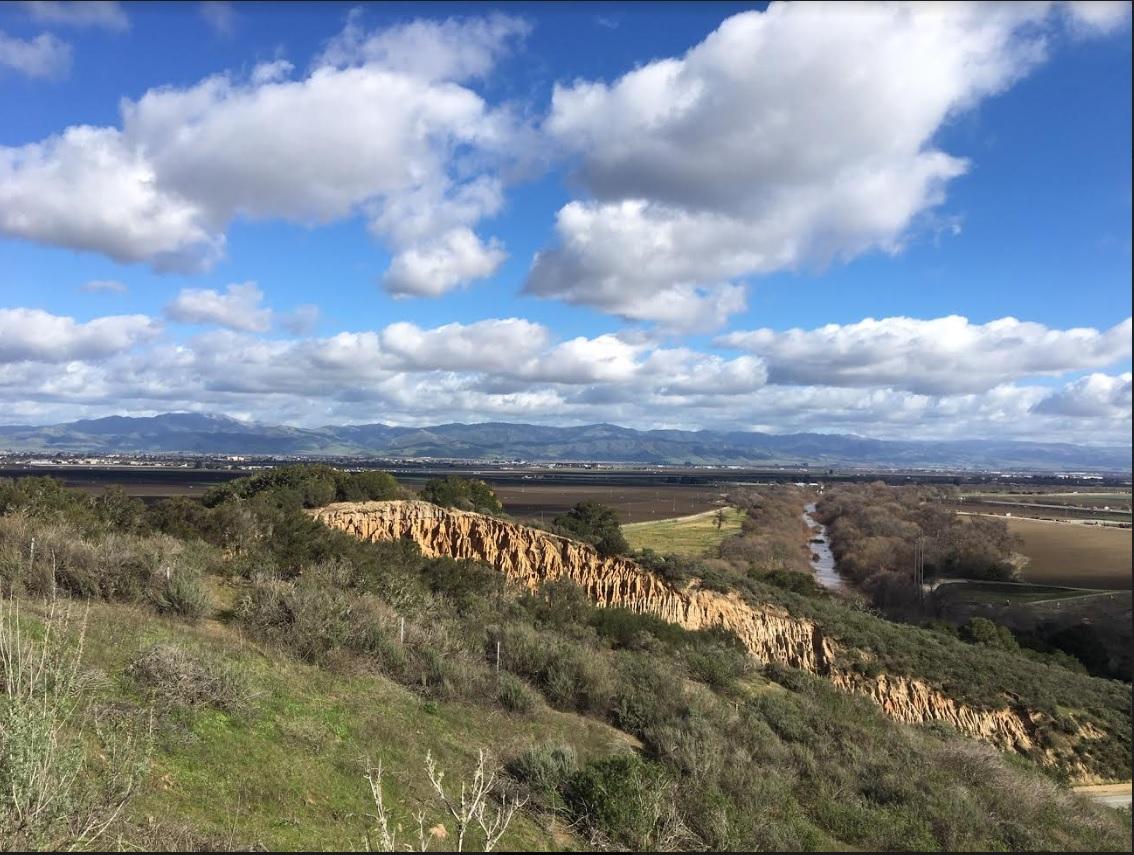 A panoramic landscape showcasing rolling hills with a rocky ridge in the foreground, lush greenery, and a winding river below. The background features a vast plain leading to distant mountains under a partly cloudy sky. Goat Trail / #41 mountain bike trail.