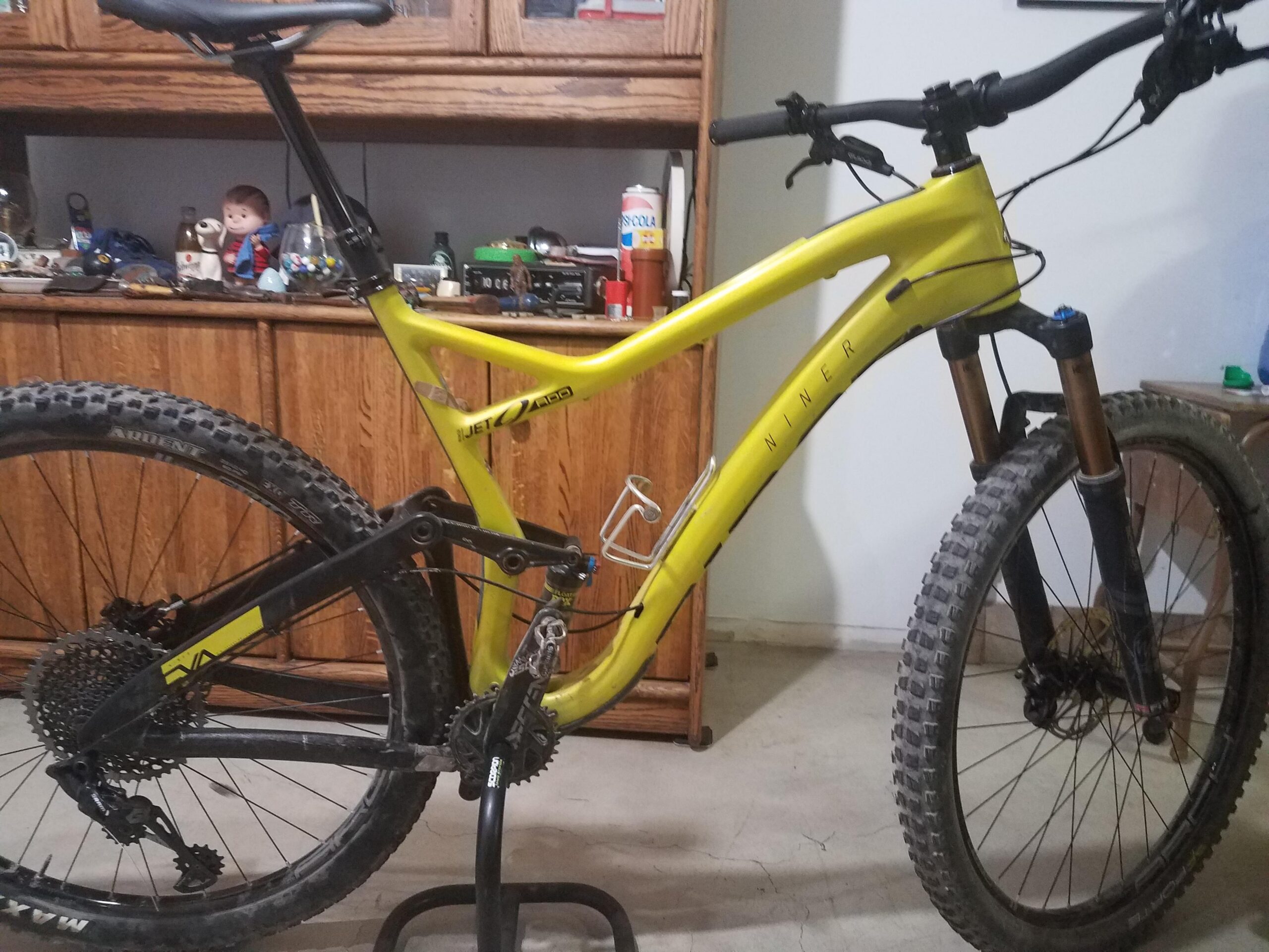 Niner Jet 9 RDO: A vibrant yellow mountain bike with a sleek frame, featuring a dropper post and front suspension. The bike is positioned on a stand, showcasing its black wheels and gear setup. In the background, there is a wooden cabinet containing various collectibles and items, adding context to the indoor setting.