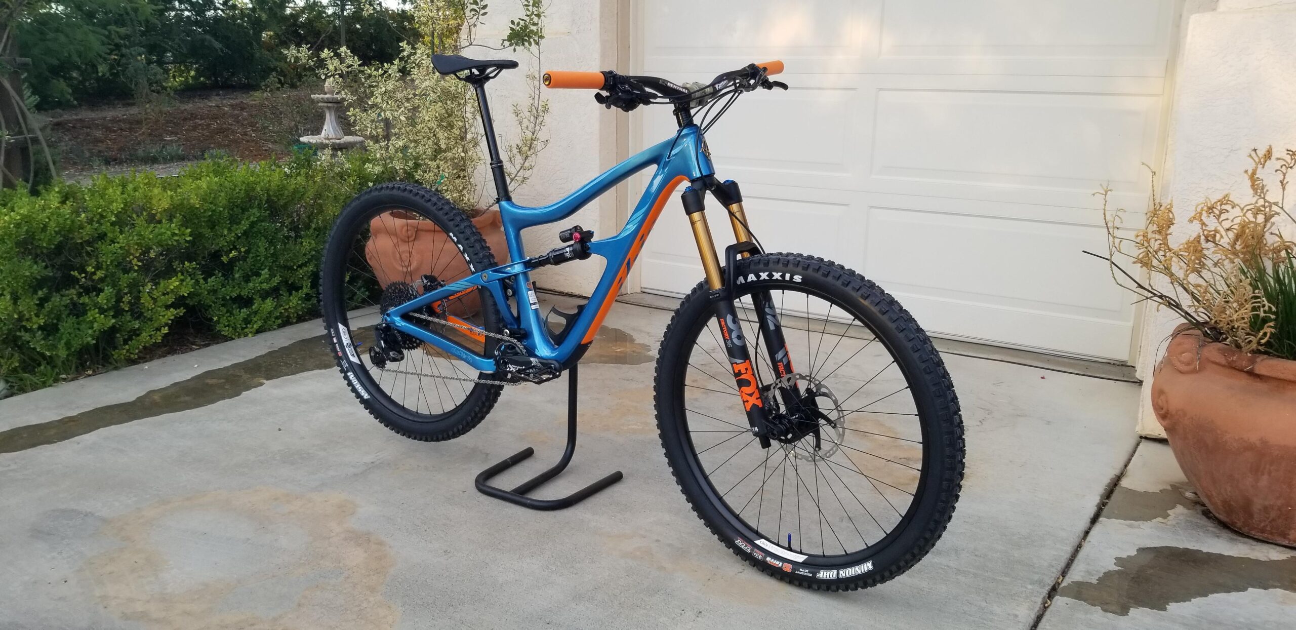 Ibis RipMo: A sleek blue mountain bike with orange accents, featuring a sturdy frame, front suspension forks, and knobby tires, is positioned on a concrete surface with a garage door in the background. The bike is resting on a stand, showcasing its design and components, surrounded by greenery and a decorative pot.