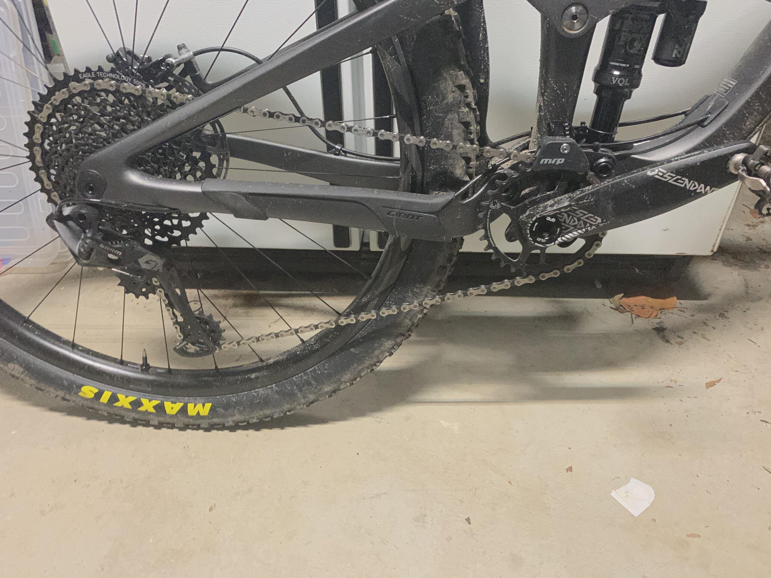 Giant Trance: Close-up image of a mountain bike's rear drivetrain, featuring a black cassette, derailleur, and chain. The bike is dirty, indicating recent use, and the tire has a visible "Maxxis" logo. The background shows a garage floor with some scattered debris.