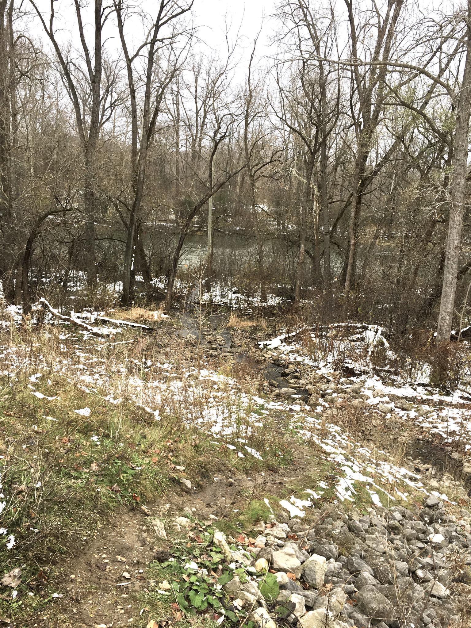 A landscape scene depicting a river surrounded by bare trees on a chilly day. The foreground features a mixture of rocky terrain and patches of grass, with remnants of snow scattered on the ground. The water is visible in the background, reflecting the overcast sky. Western University trails mountain bike trail.