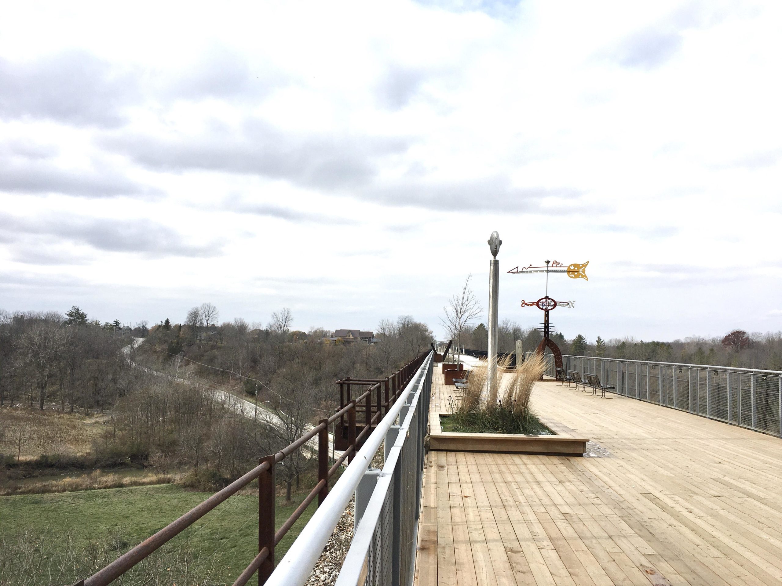 A wooden walkway with a railing, overlooking a landscape of trees and a winding road. The sky is overcast with gray clouds. Decorative weather vanes are visible along the path, adding a whimsical touch to the scene. Union station back woods mountain bike trail.