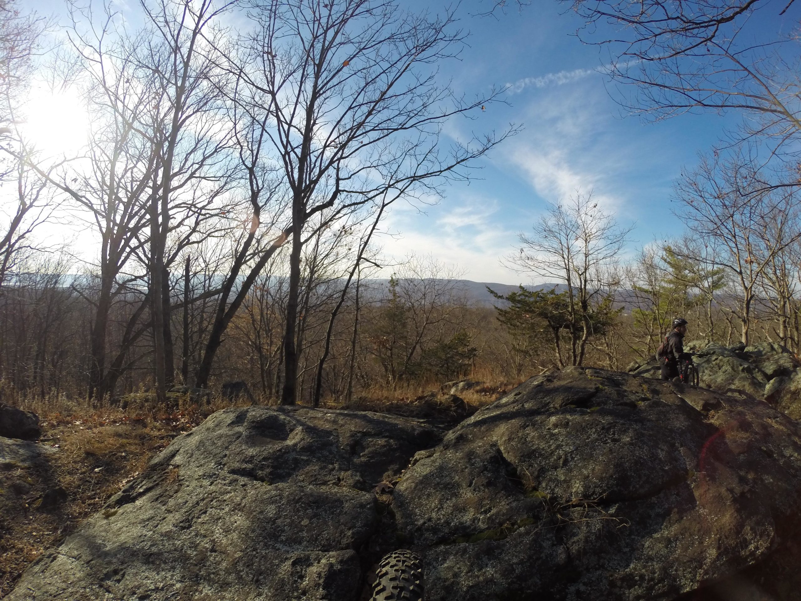 A scenic view of a mountainous landscape in early spring, featuring bare trees and large rocks. A cyclist stands near the rocks, overlooking the valley below with trees and mountains in the background under a bright blue sky dotted with clouds. The sun shines through the branches, casting a warm light on the scene. Blue Mountain Reservation mountain bike trail.