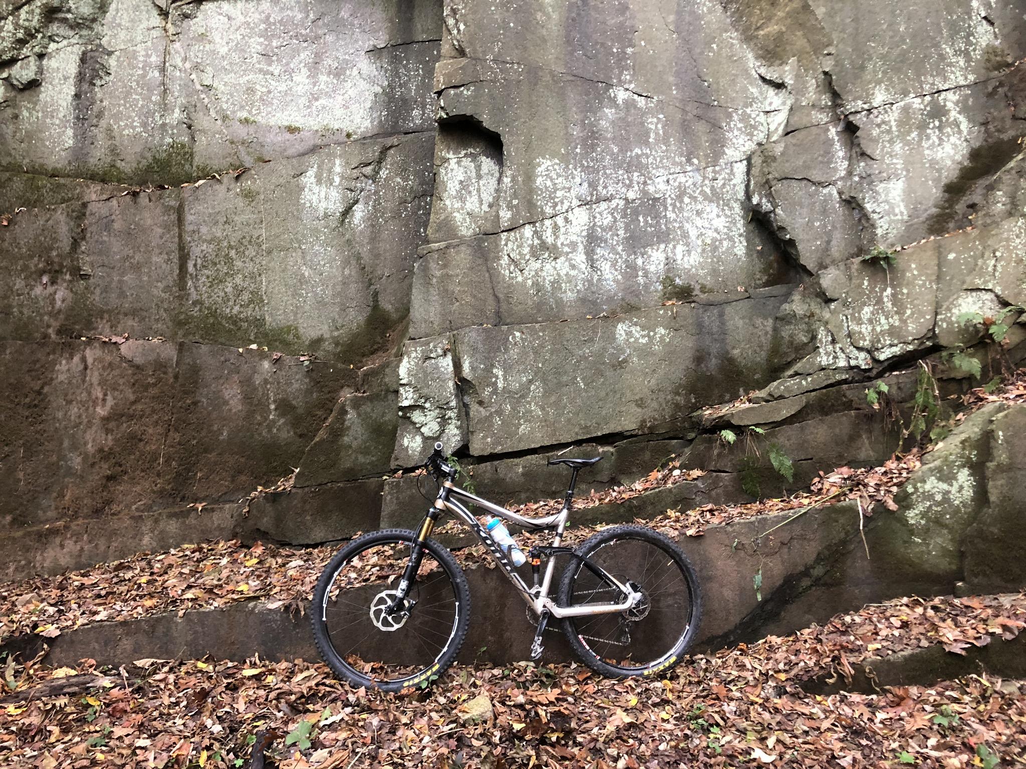A mountain bike leaning against a large, textured rock wall, surrounded by a layer of dry leaves. The scene is set in a natural outdoor environment, showcasing earthy tones and vegetation. Susquehanna mountain bike trail.
