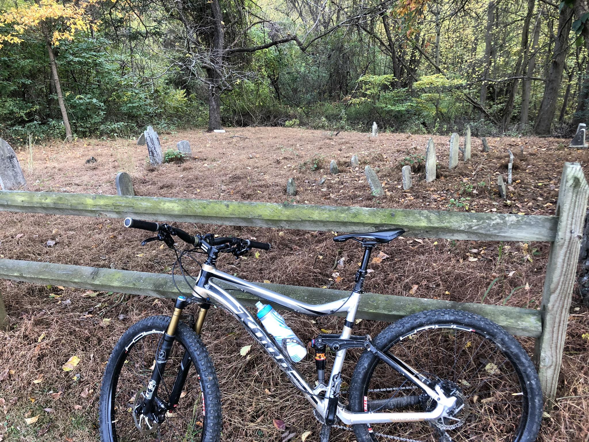 A mountain bike resting against a wooden fence, overlooking a small graveyard with several gravestones among overgrown grass and foliage. Trees with autumn foliage surround the scene, creating a peaceful, natural atmosphere. Susquehanna mountain bike trail.