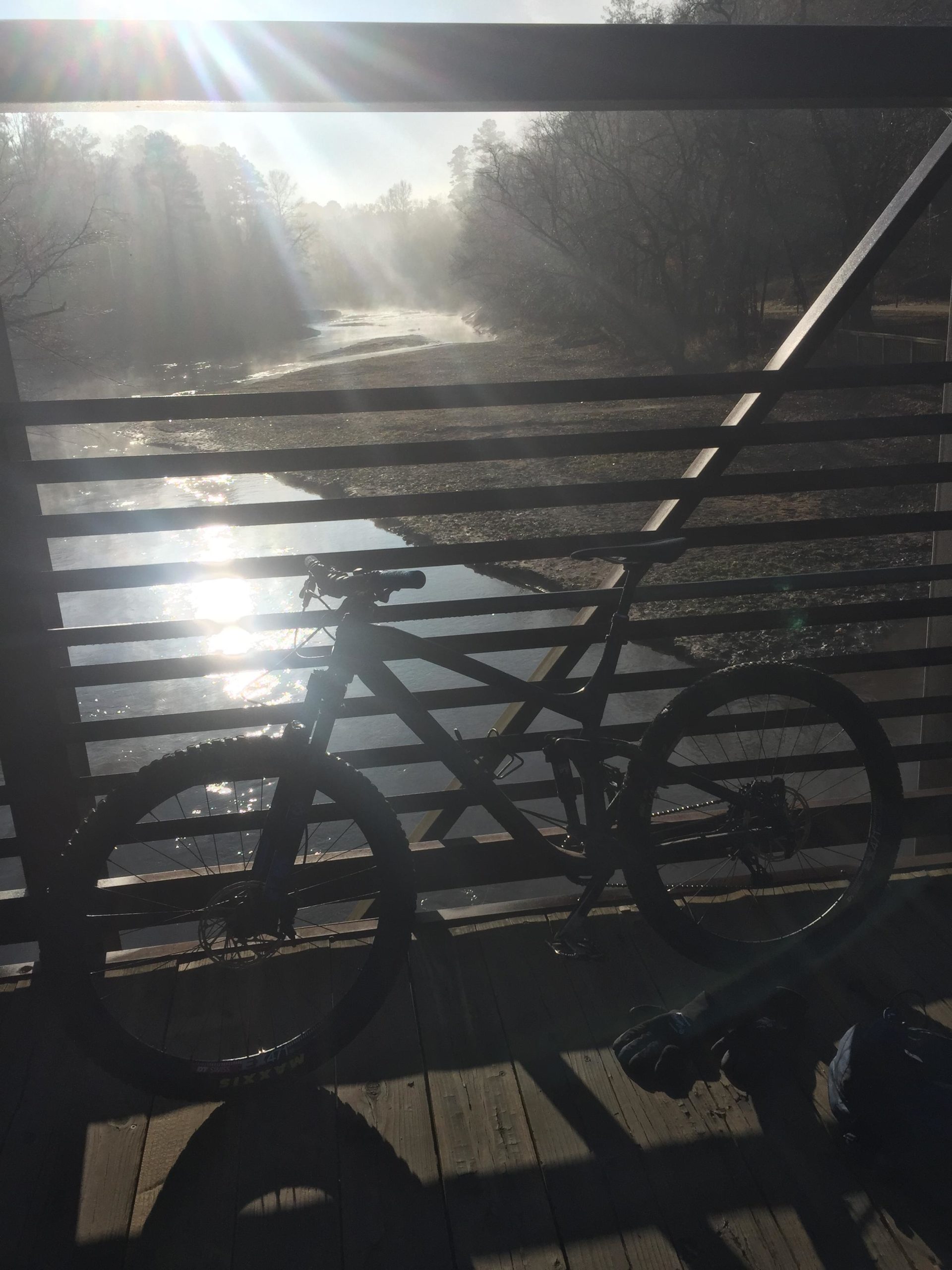 A mountain bike rests on a wooden bridge overlooking a shimmering river, with sunlight streaming through the trees and mist rising from the water. The scene captures a serene and tranquil outdoor setting. Taylor Randahl Memorial Mountain Bike Trails At Olde Rope Mill Park mountain bike trail.