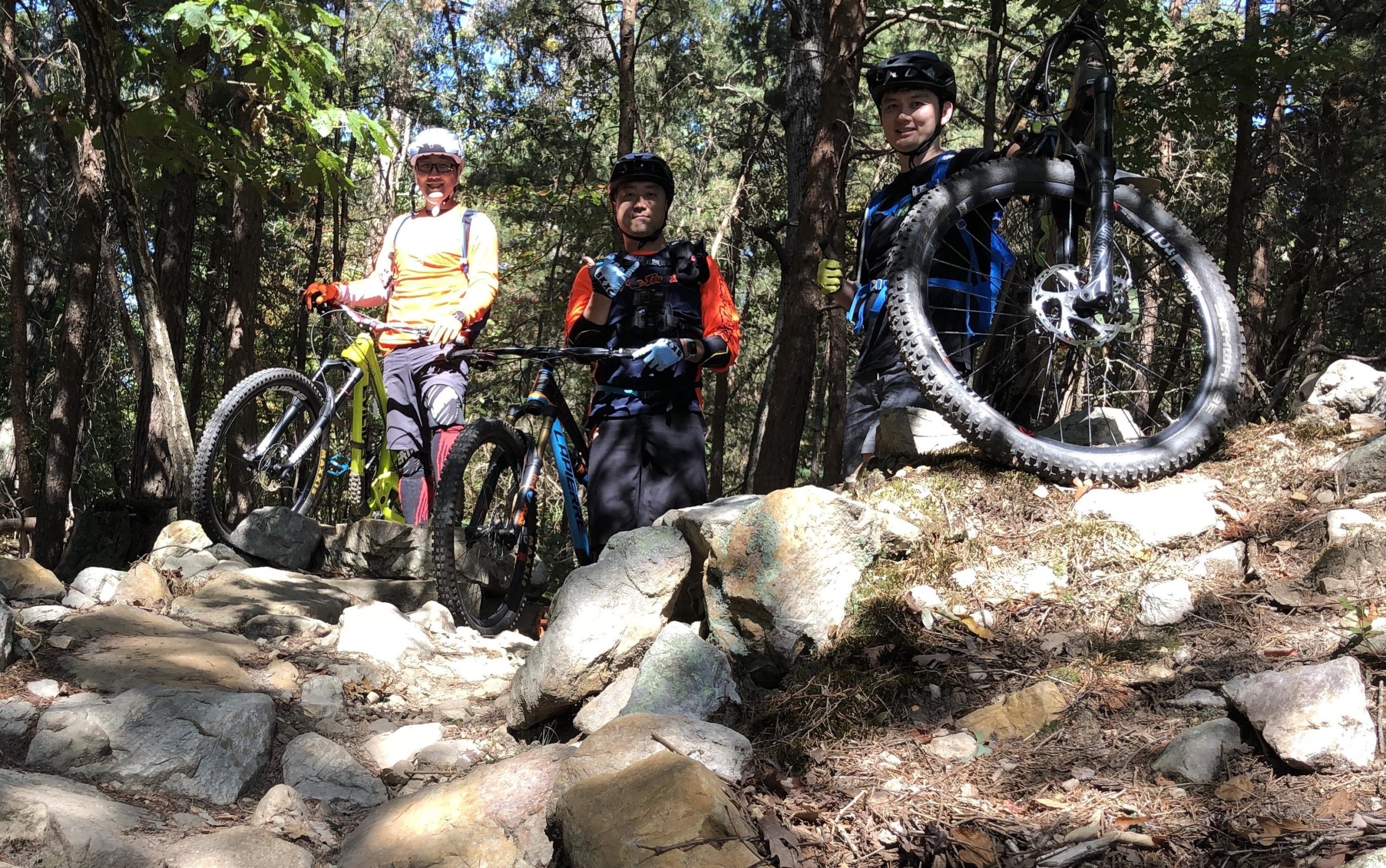 Three mountain bikers posing on a rocky trail in a forested area, with two riders holding their bikes and one with a bike resting on rocks. They are wearing helmets and colorful cycling attire, surrounded by trees and sunlight filtering through the leaves. Massanutten Western Slope mountain bike trail.