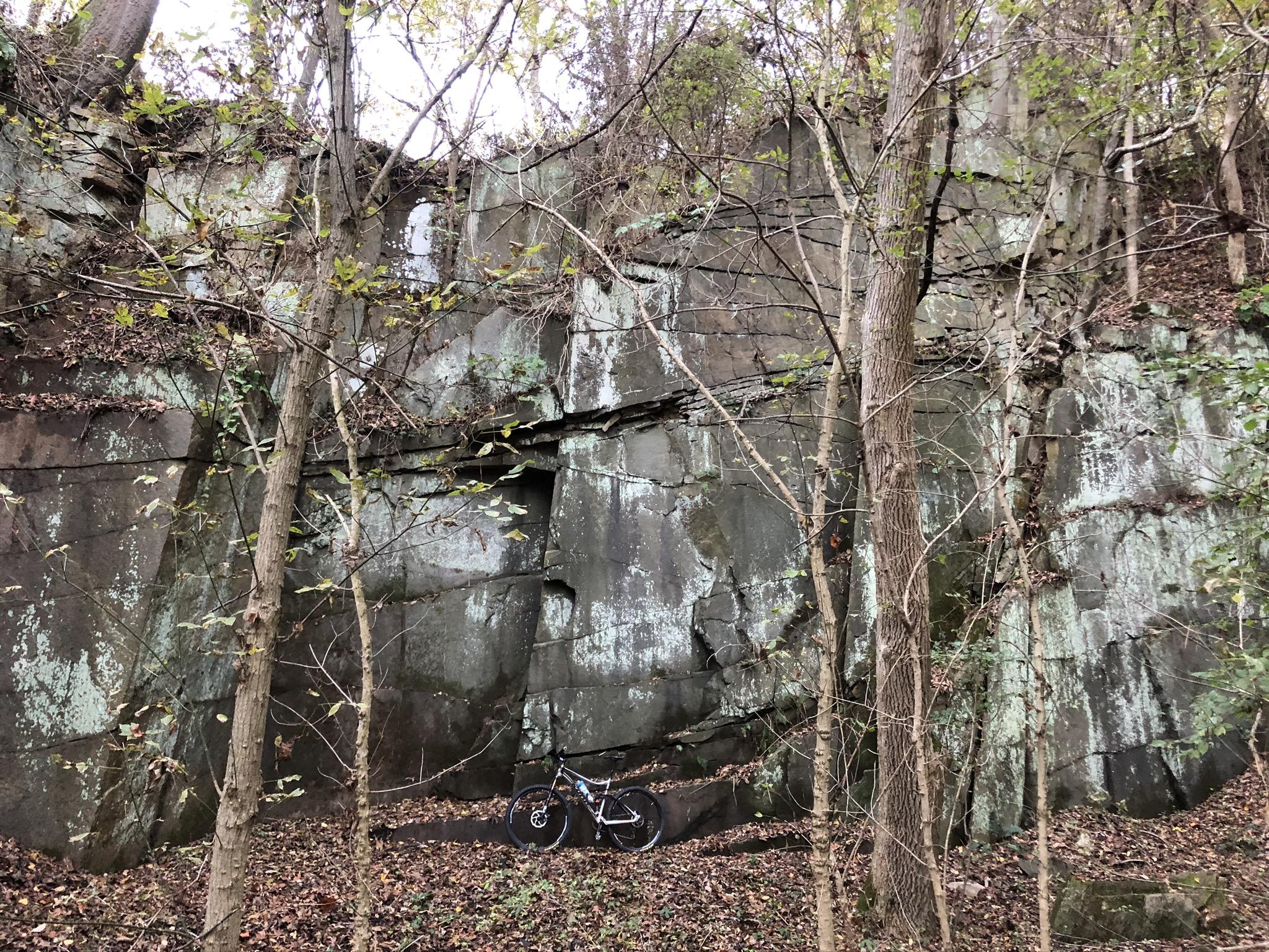 A rocky cliff covered with patches of moss, surrounded by trees with sparse leaves. A mountain bike rests on the forest floor amidst scattered fallen leaves. The scene captures a peaceful moment in a wooded area. Susquehanna mountain bike trail.