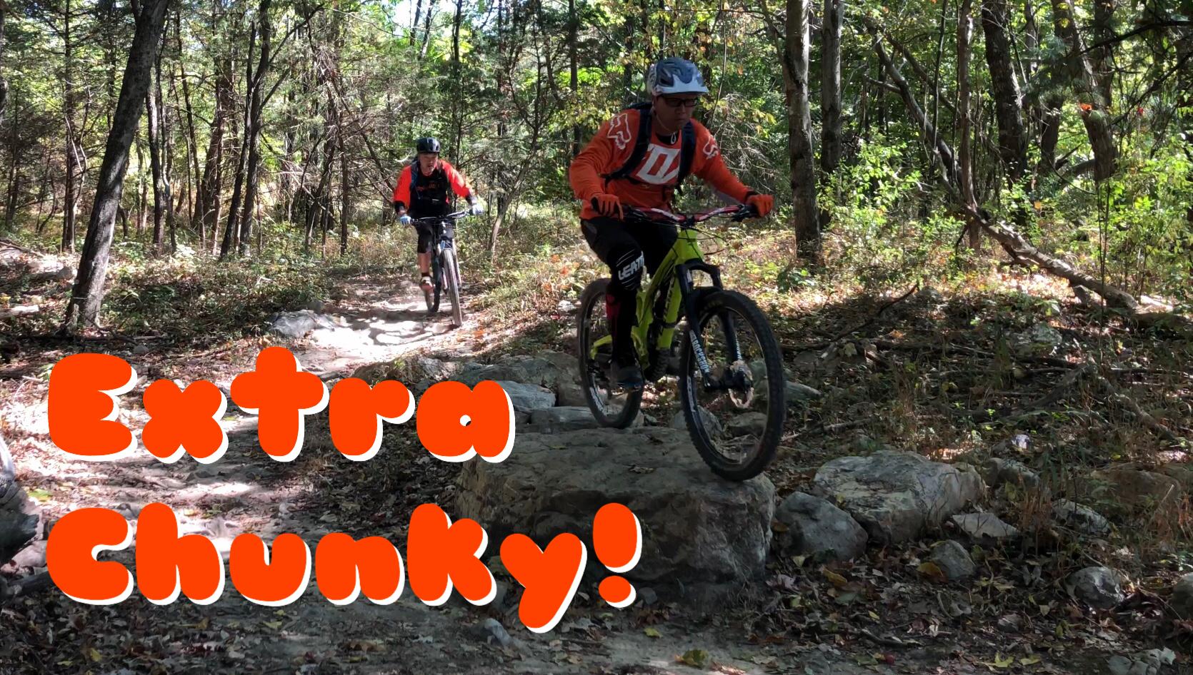 Two mountain bikers navigate a rocky trail in a forested area. One cyclist is riding over a large rock, while the other follows behind on the path. The scene conveys an adventurous outdoor atmosphere, with a focus on the technical challenges of mountain biking, highlighted by the phrase "Extra Chunky!" in bold letters at the bottom. The surrounding trees and foliage suggest a vibrant autumn backdrop. Massanutten Western Slope mountain bike trail.