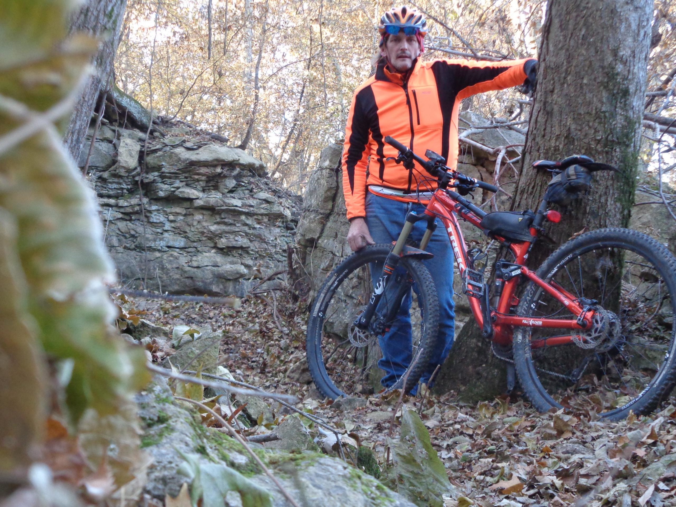 A person in an orange and black cycling jacket stands beside a red mountain bike, leaning against a tree in a wooded area. The background features rocky terrain and fallen leaves, indicating an outdoor setting perfect for biking. Big Bull Creek mountain bike trail.