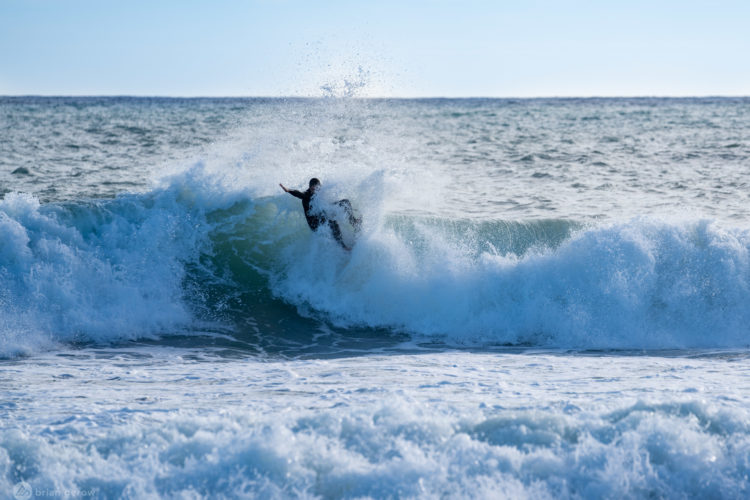 A surfer riding a large wave, creating a splash of water, with a clear blue sky and ocean in the background.