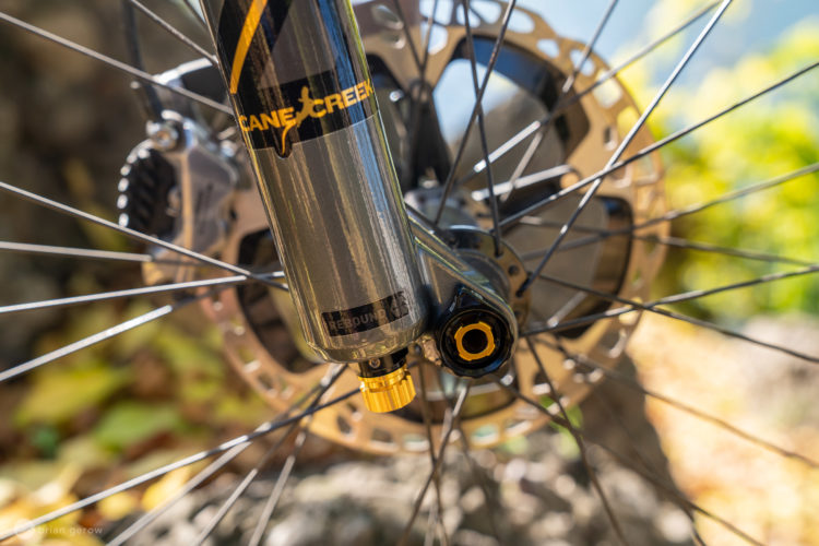 Close-up of a bicycle suspension fork with visible branding from Cane Creek. The image highlights the rebound adjustment knob at the bottom of the fork leg, with wheel spokes and a bicycle disc brake rotor in the background. The setting appears to be outdoors with a blurred natural backdrop.