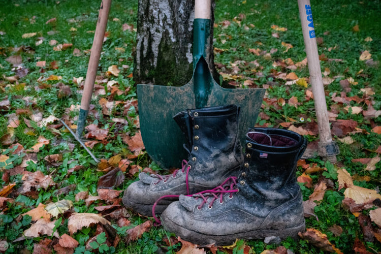 A pair of muddy work boots placed on the ground among fallen autumn leaves, with a green shovel and a garden hoe leaning against a tree in the background. The scene captures a relaxed outdoor gardening setting.