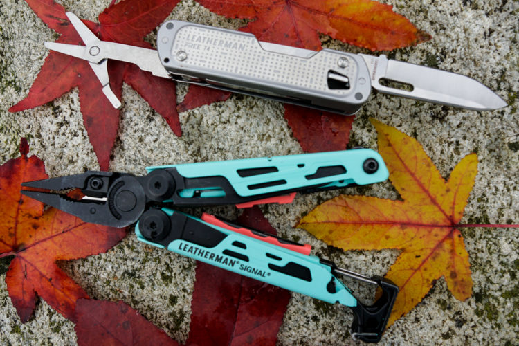 Two multi-tools are positioned on a textured surface adorned with colorful autumn leaves. The top tool is a silver Leatherman with a pair of scissors and a knife blade visible. The bottom tool is a teal and black Leatherman Signal with pliers displayed. The vibrant red and yellow leaves around them create a warm, seasonal backdrop.