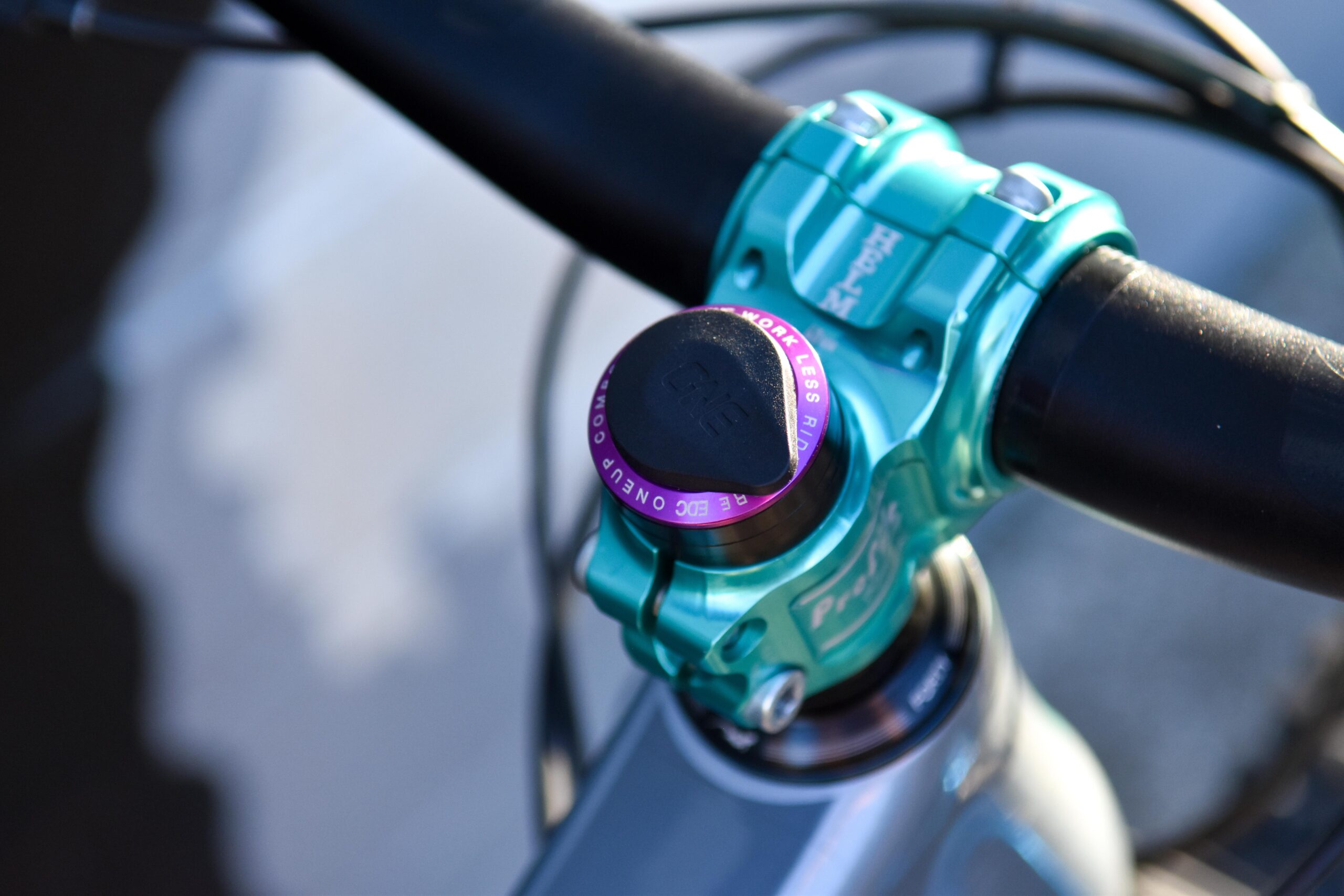 Kona Process 153 CR/DL: Close-up of a bicycle's handlebars showcasing a teal colored stem with a distinctive purple and black compression cap. The image focuses on the intricate design and detailing of the stem, emphasizing the textured grip and components.