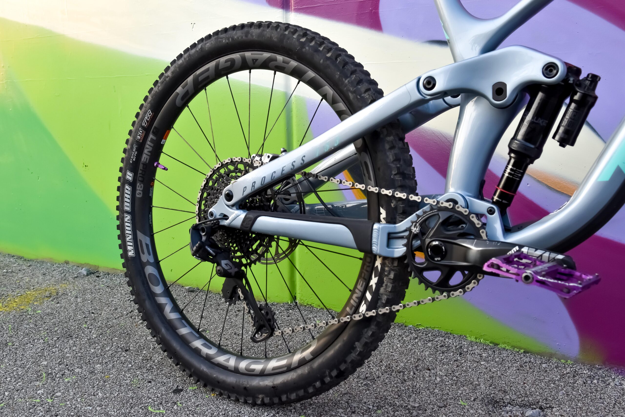 Kona Process 153 CR/DL: Close-up of the rear section of a mountain bike featuring a sleek, light blue frame and black tires. The bike is positioned against a vibrant graffiti backdrop with green and purple hues. Key components visible include the rear suspension, chain, and a purple pedal, highlighting its design and detailing.