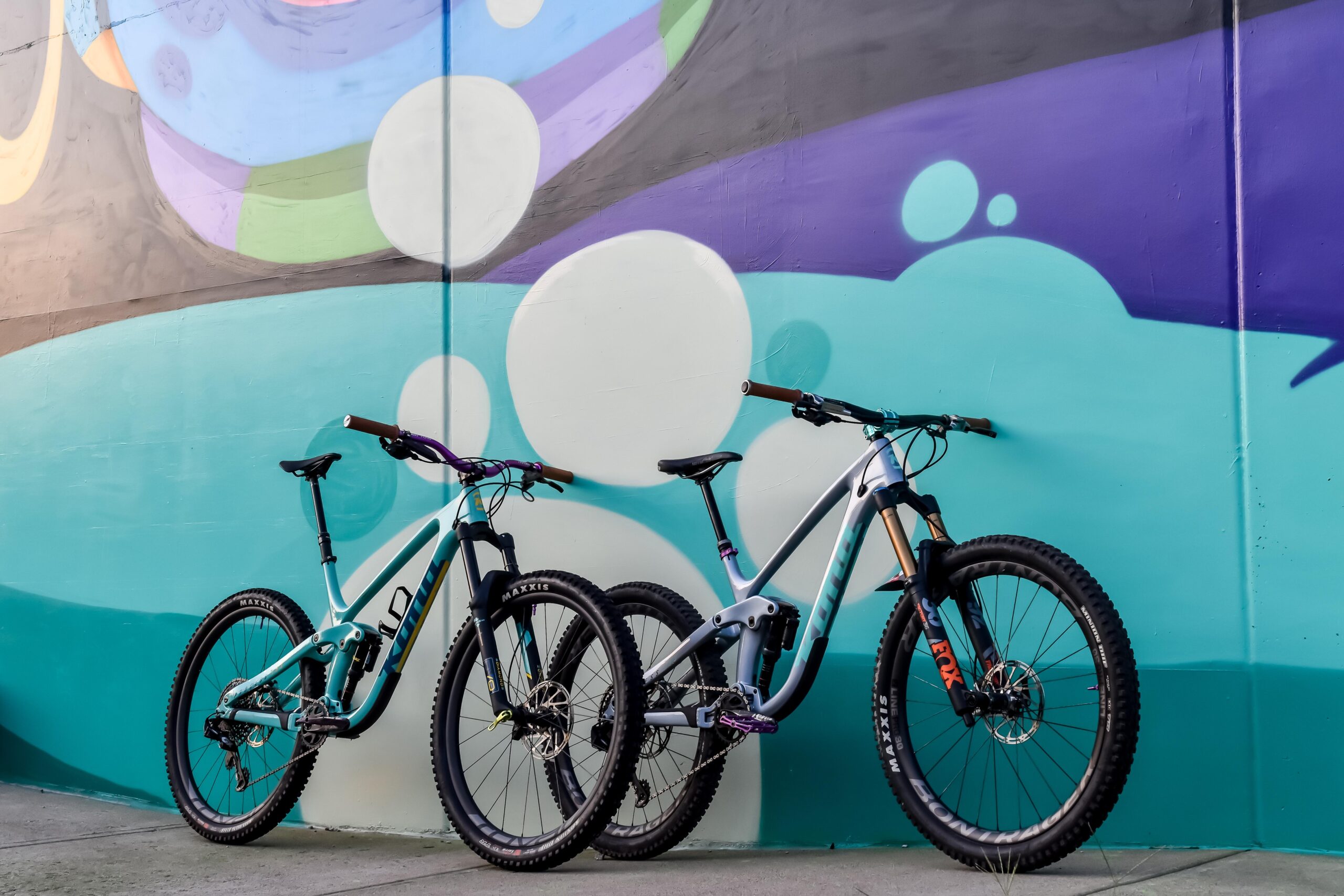 Kona Process 153 CR/DL: Two mountain bikes are parked side by side in front of a vibrant mural featuring shades of blue, purple, and white. The bikes have distinctive frames and thick tires, suggesting they are designed for off-road cycling. The mural serves as a colorful backdrop to the scene.
