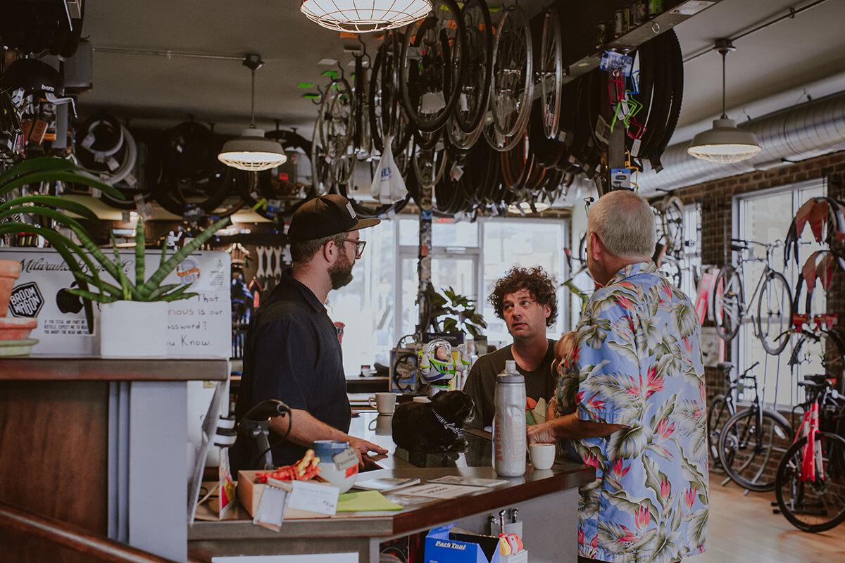 A bike shop interior featuring three men engaged in conversation at the counter. Bicycles and cycling equipment are visible on the walls, and a green plant sits on the counter. One man wears a baseball cap and glasses, while another has curly hair. A third man in a floral shirt stands with a drink in hand. The scene conveys a lively atmosphere in a local bike shop.