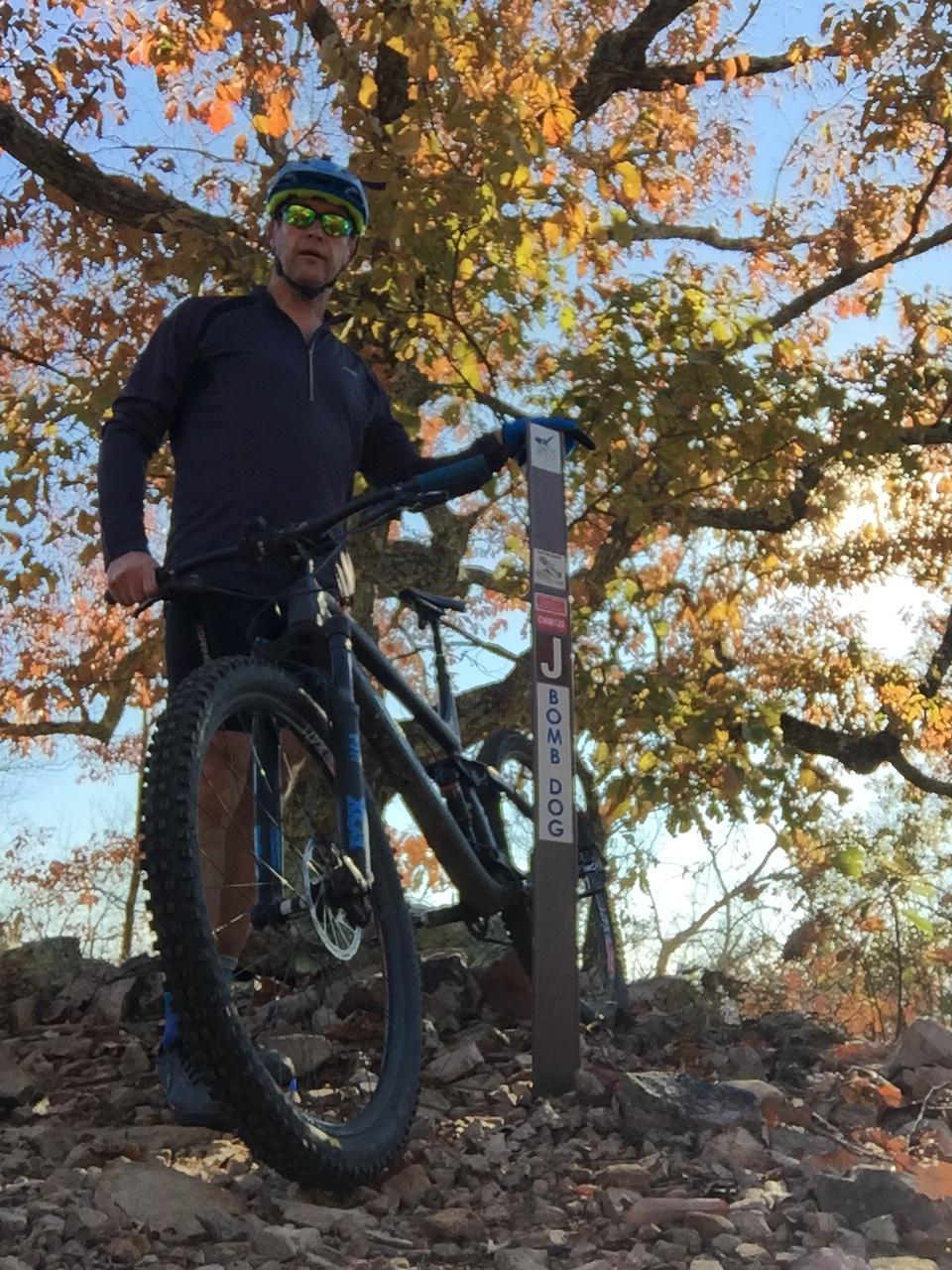 A mountain biker wearing a blue helmet and sunglasses stands next to his bike on a rocky trail, surrounded by trees with colorful autumn foliage. A trail sign labeled "J BOMBDOG" is visible in the foreground. Coldwater Mountain mountain bike trail.
