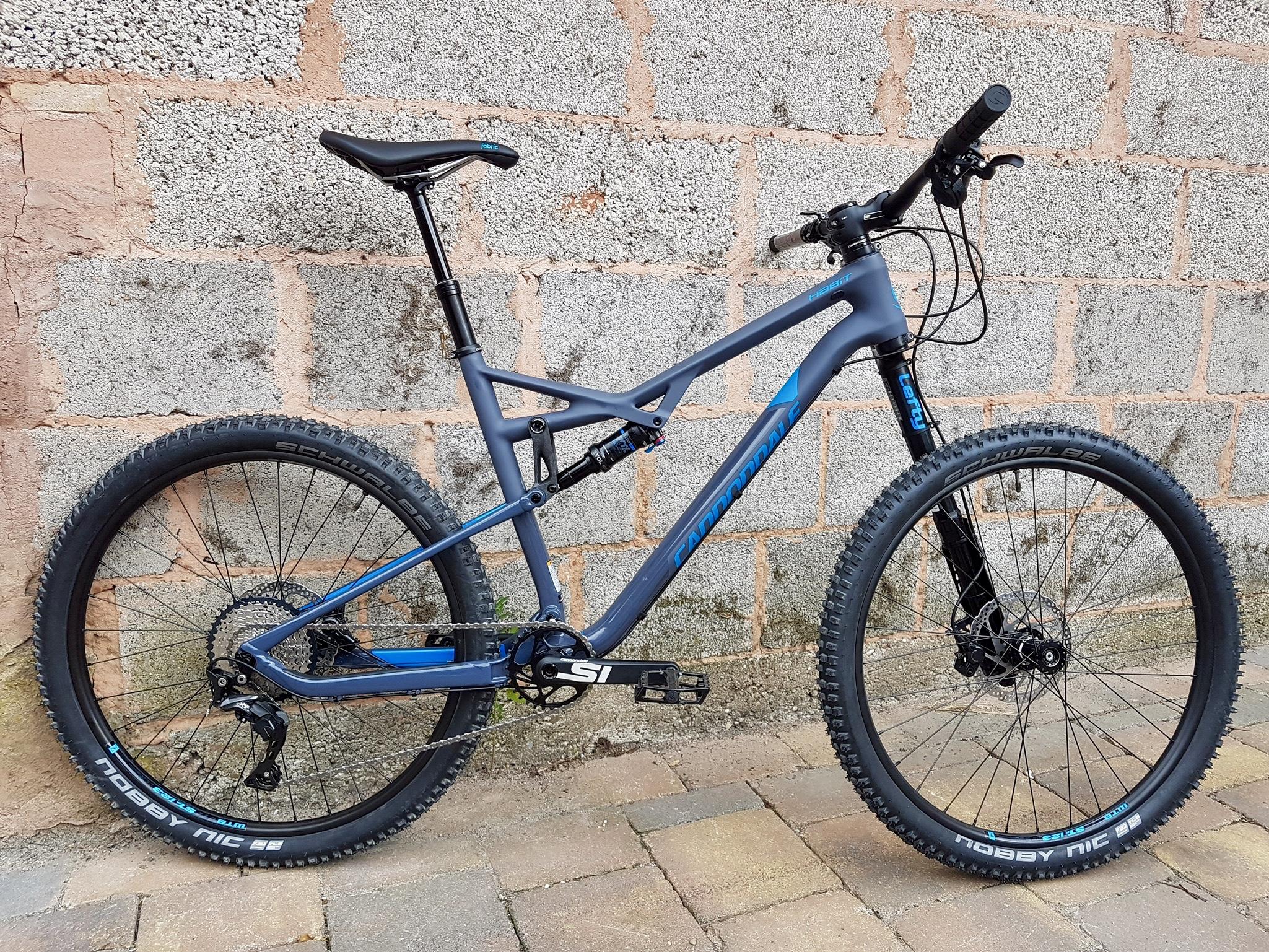 A modern mountain bike with a gray and blue frame, featuring knobby tires, hydraulic disc brakes, and a prominent suspension system, leaning against a textured concrete wall. The bike's components include a black saddle, adjustable seat post, and a visible gear system on the rear wheel. The setting includes a paved surface and an uneven wall backdrop. melkerei 45Km loop mountain bike trail.