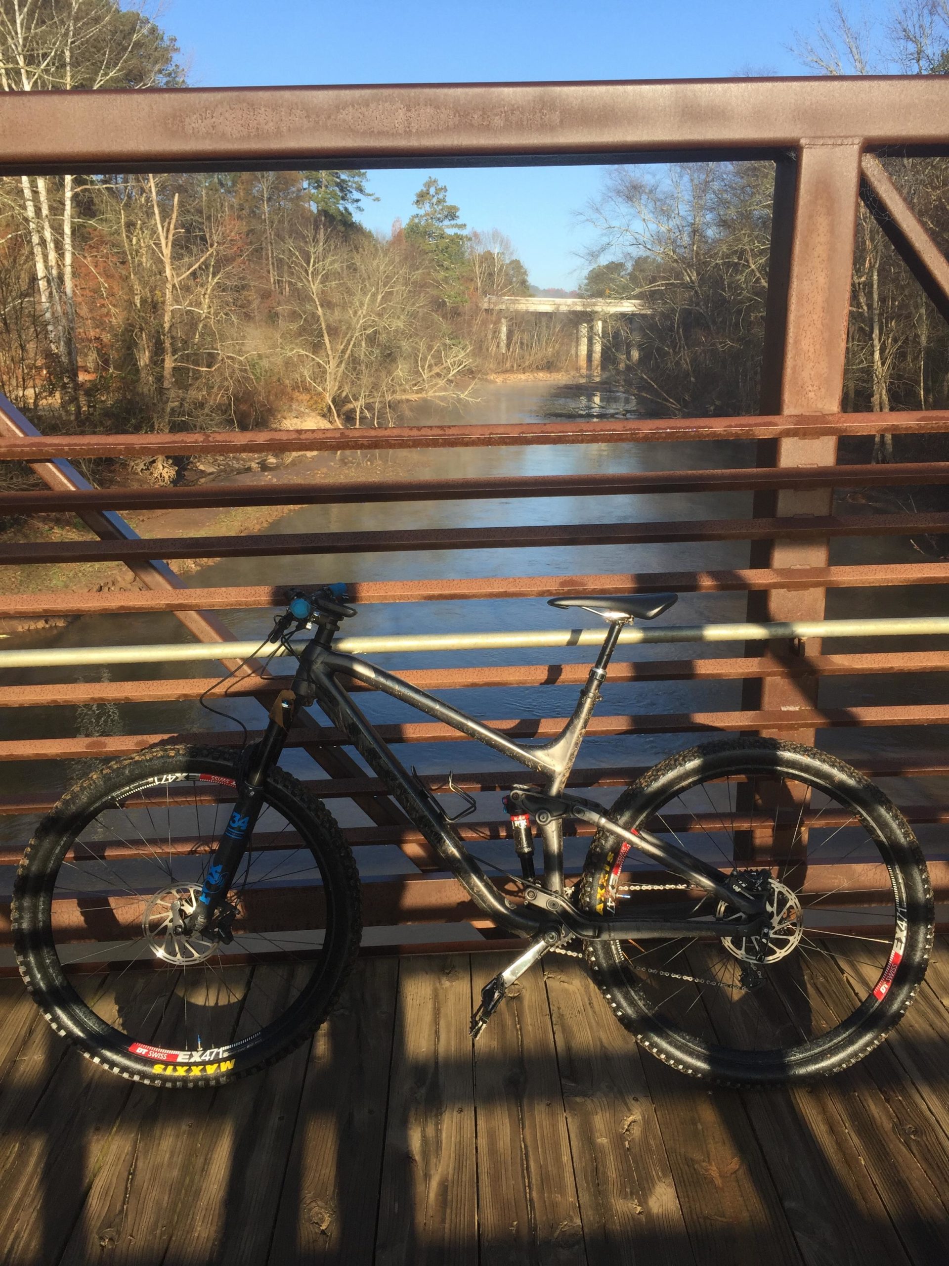 A mountain bike is positioned on a wooden bridge overlooking a calm river. The background features trees with bare branches, and a concrete bridge is visible in the distance. The sky is clear and blue, indicating a sunny day. Taylor Randahl Memorial Mountain Bike Trails At Olde Rope Mill Park mountain bike trail.