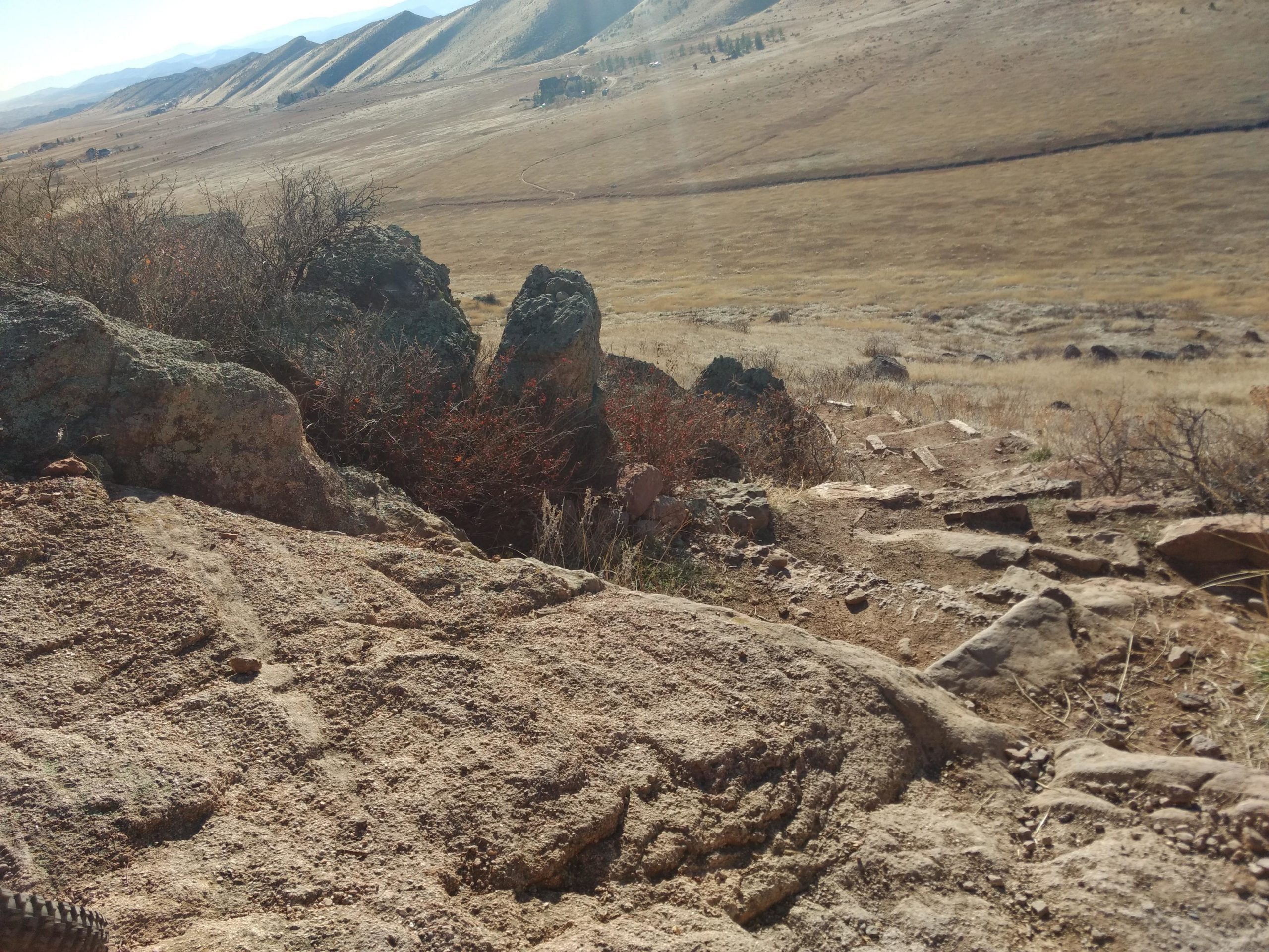 A rocky landscape with sparse vegetation, set against rolling hills and distant mountains. The foreground features textured rocks and some red bushes, while the background shows a grassy area leading into the horizon. Coyote Ridge mountain bike trail.